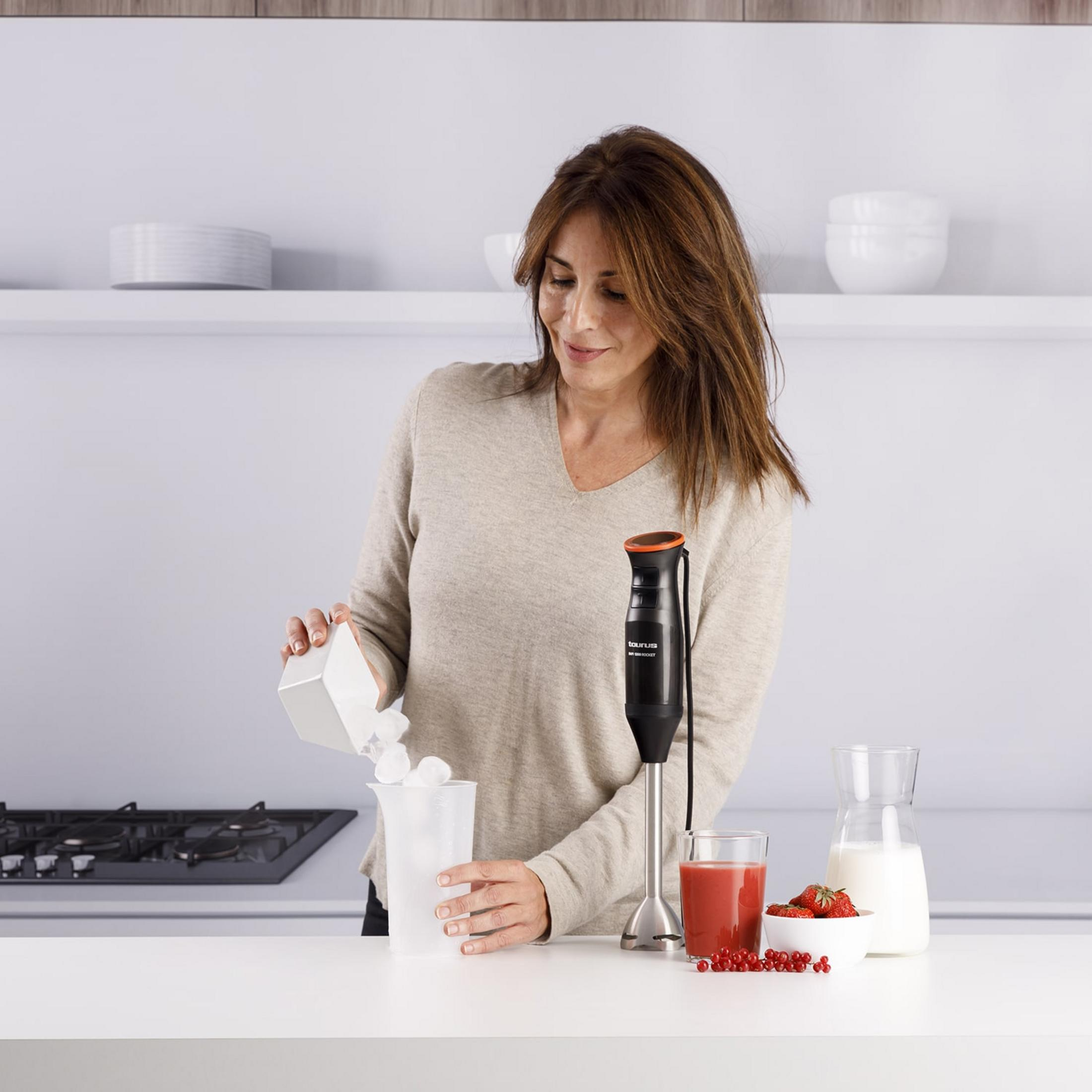 Donna versa ghiaccio in un bicchiere accanto a un frullatore, succo e bacche.