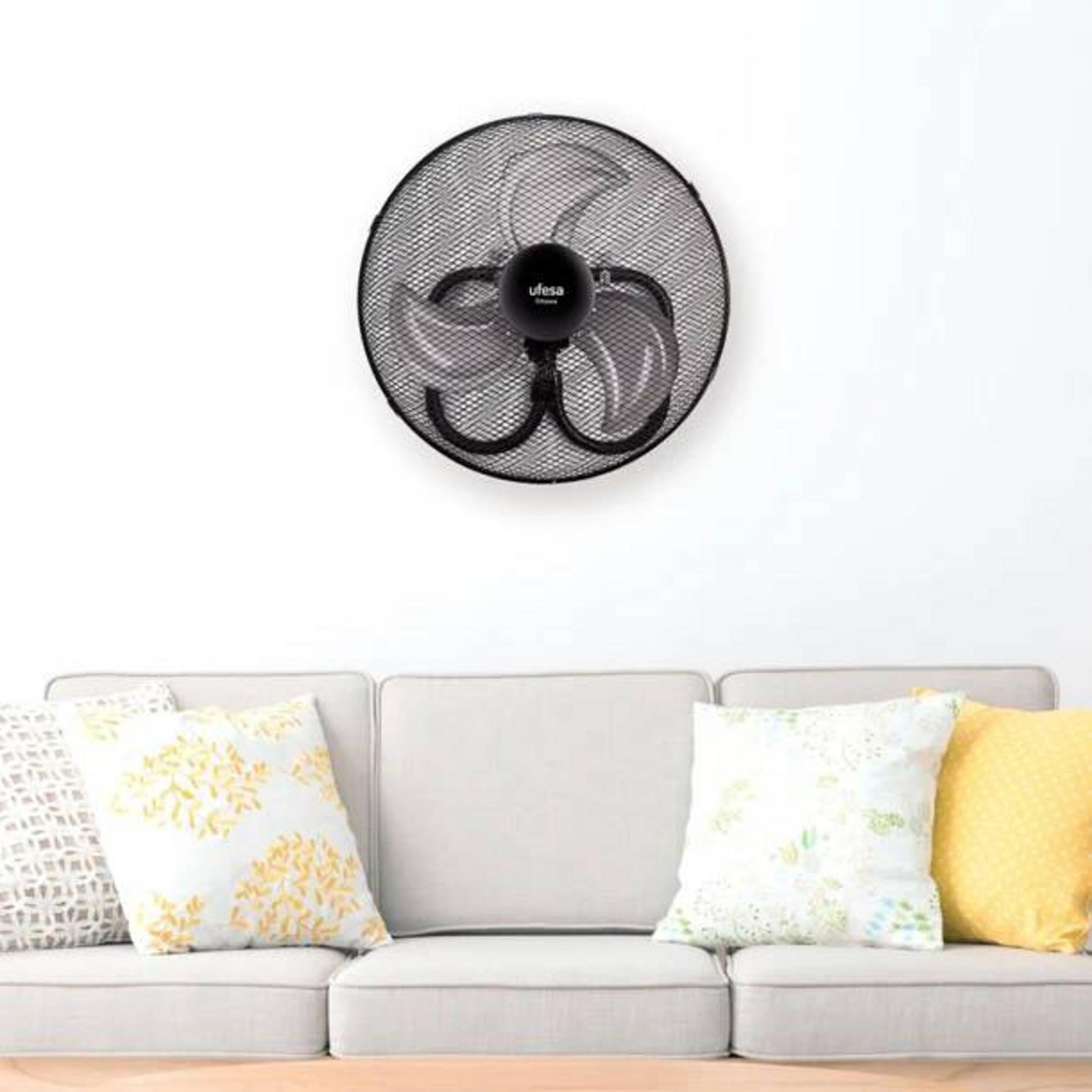 Un ventilador negro de pared cuelga sobre un sofá blanco con cojines coloridos.