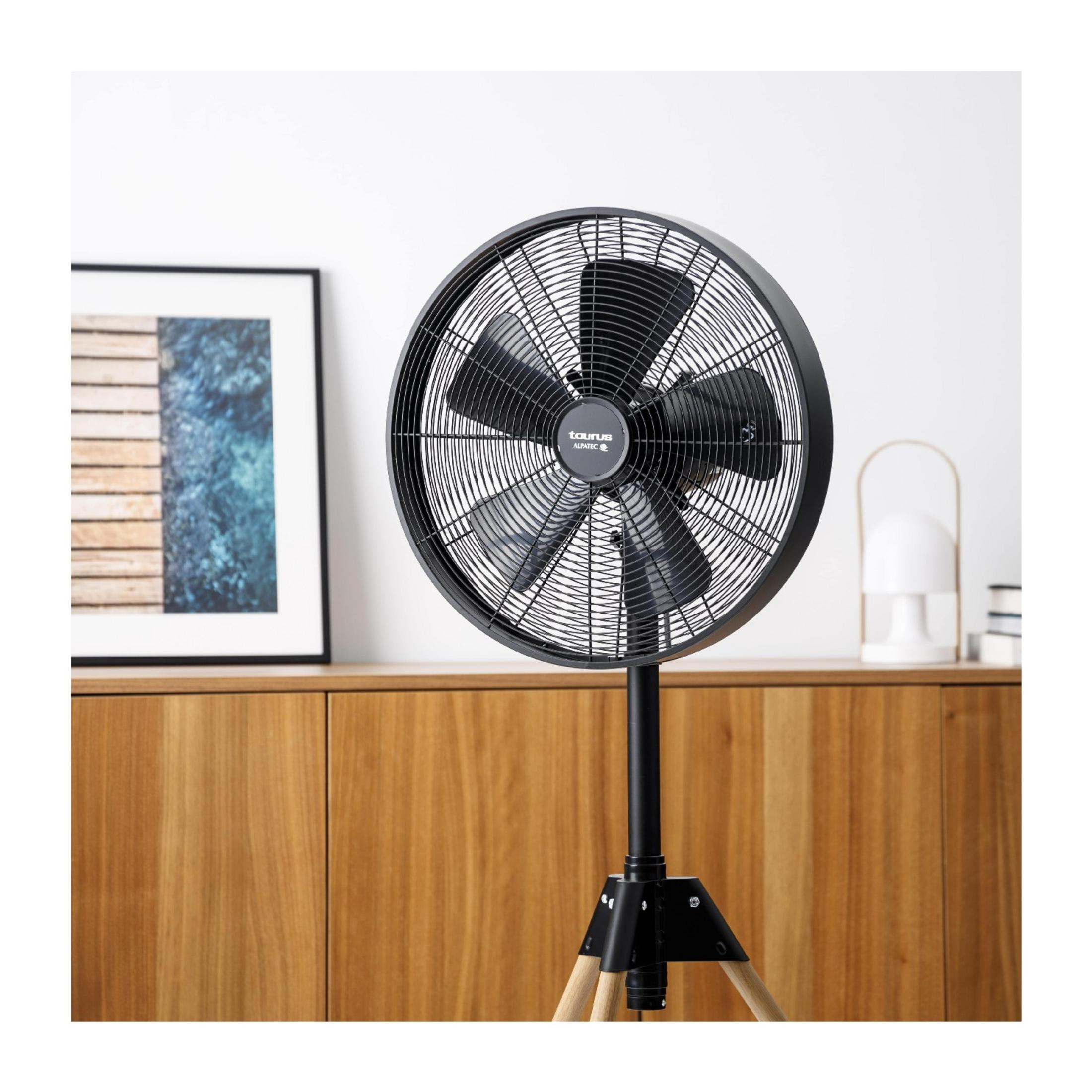 Un ventilador de pie negro con detalles de madera está frente a un mueble de madera con un cuadro.