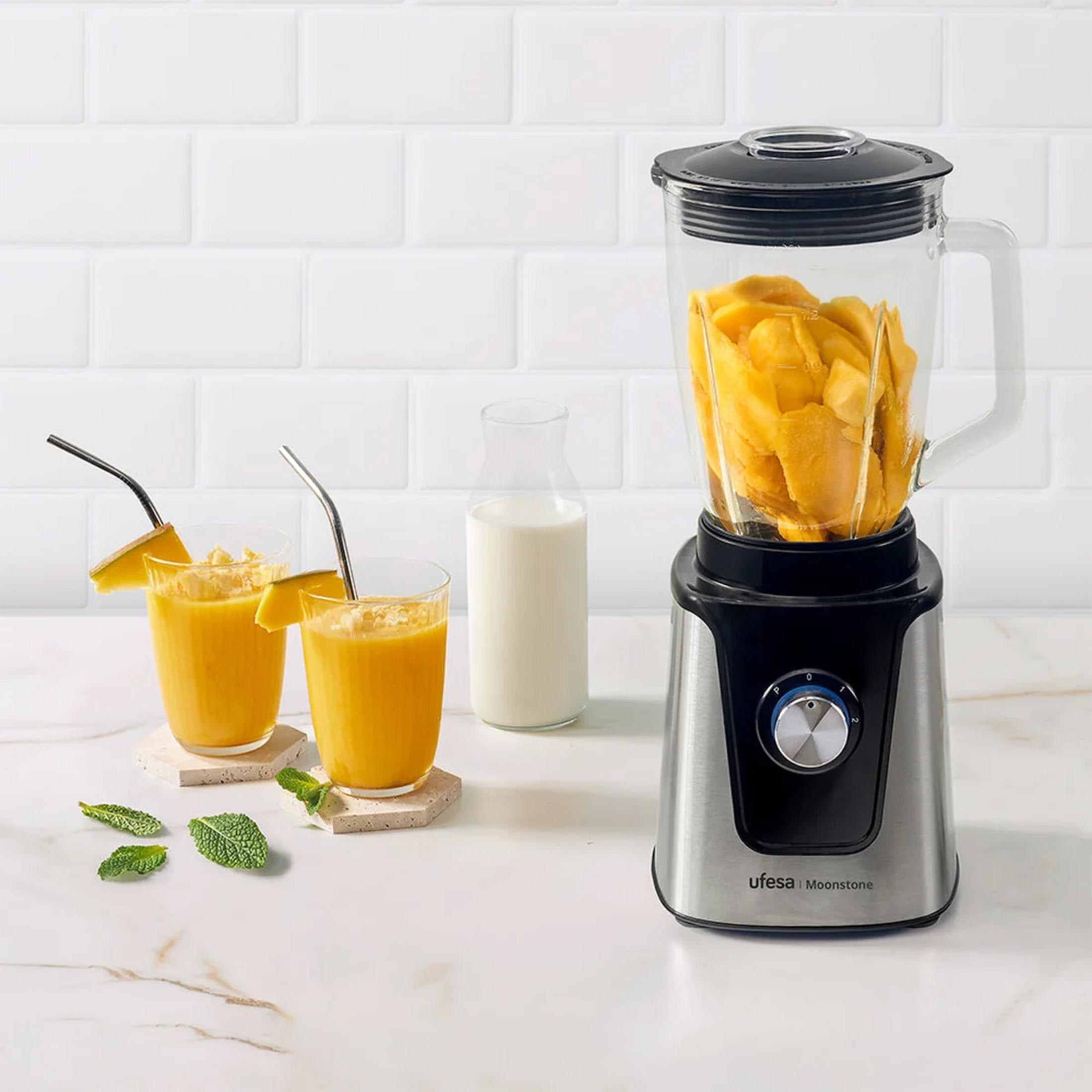 Un frullatore riempito di mango, accanto a due bicchieri di succo e una bottiglia di latte.