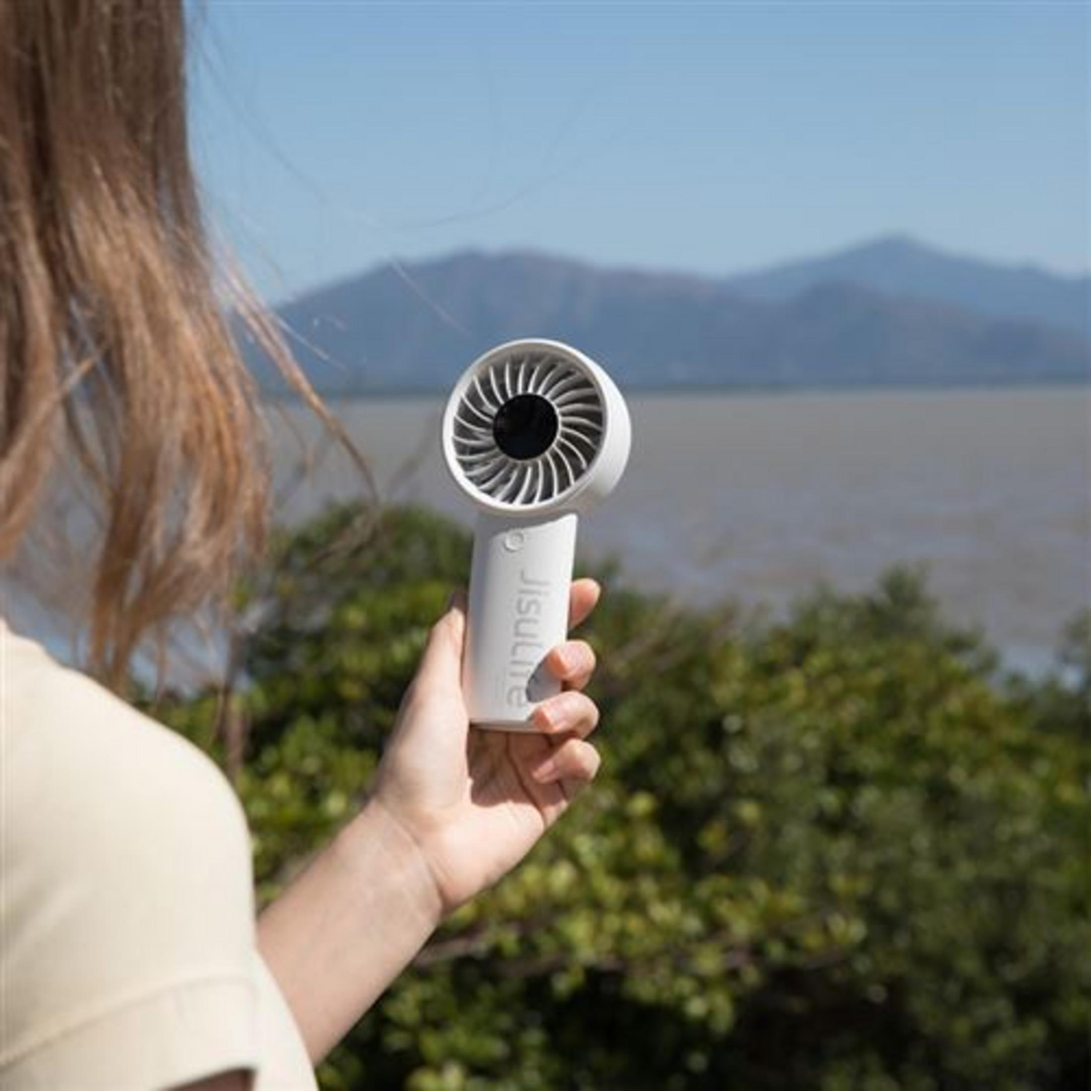 Una mano bianca tiene un piccolo ventilatore davanti a un paesaggio con montagne e acqua.