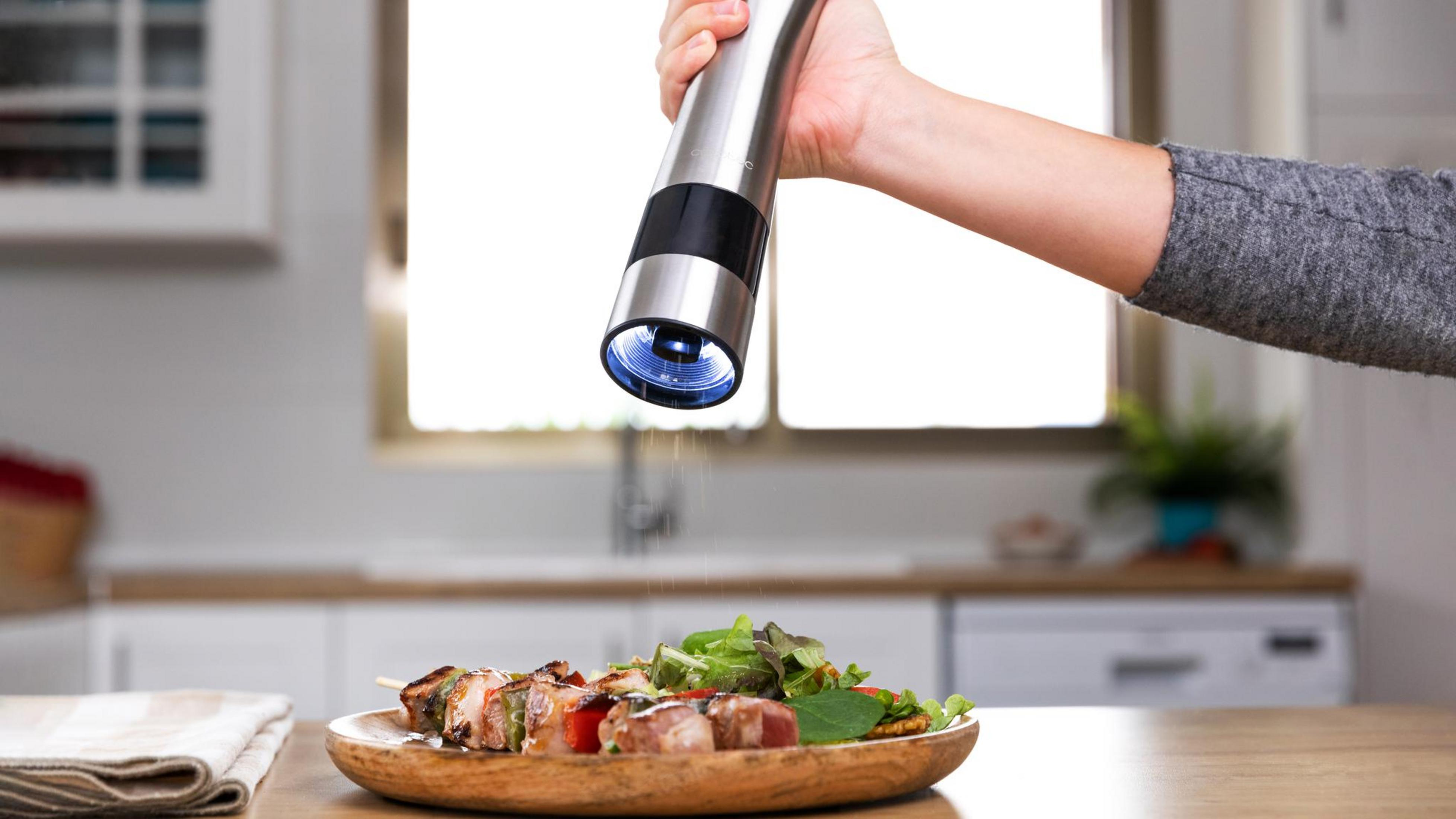 A hand uses an electric grinder over a plate of meat skewers and salad.