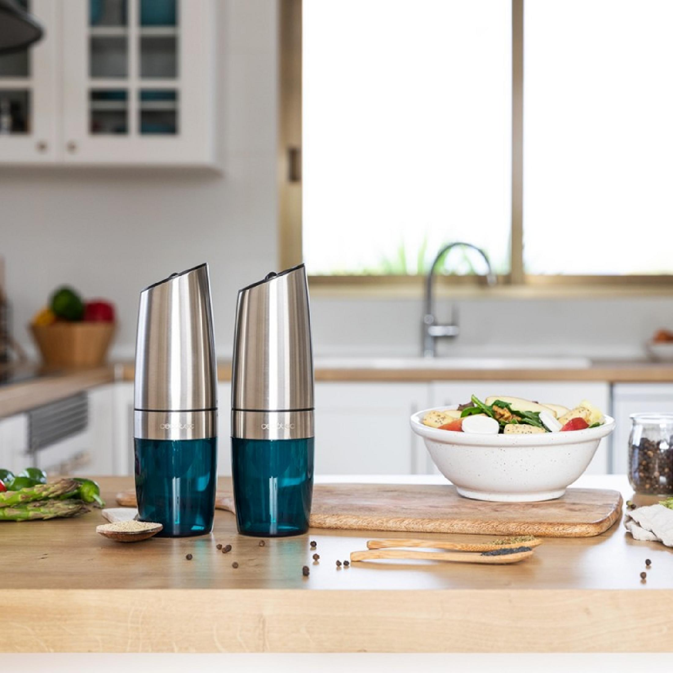 Two stainless steel salt and pepper grinders with blue glass sit on a wooden table next to a salad bowl.