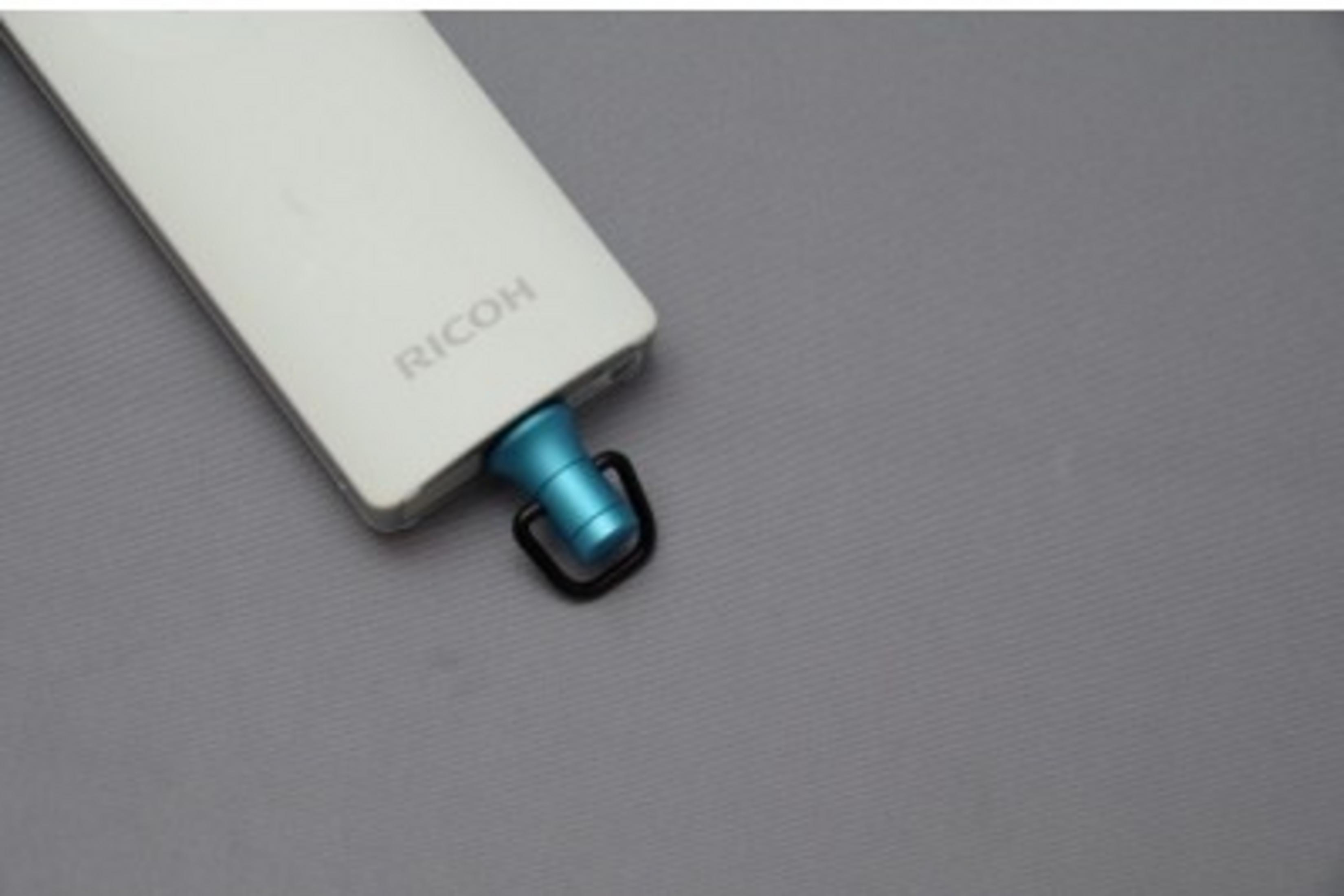 Białe urządzenie Ricoh z niebieskim dodatkiem i czarnym pierścieniem na szarym tle.