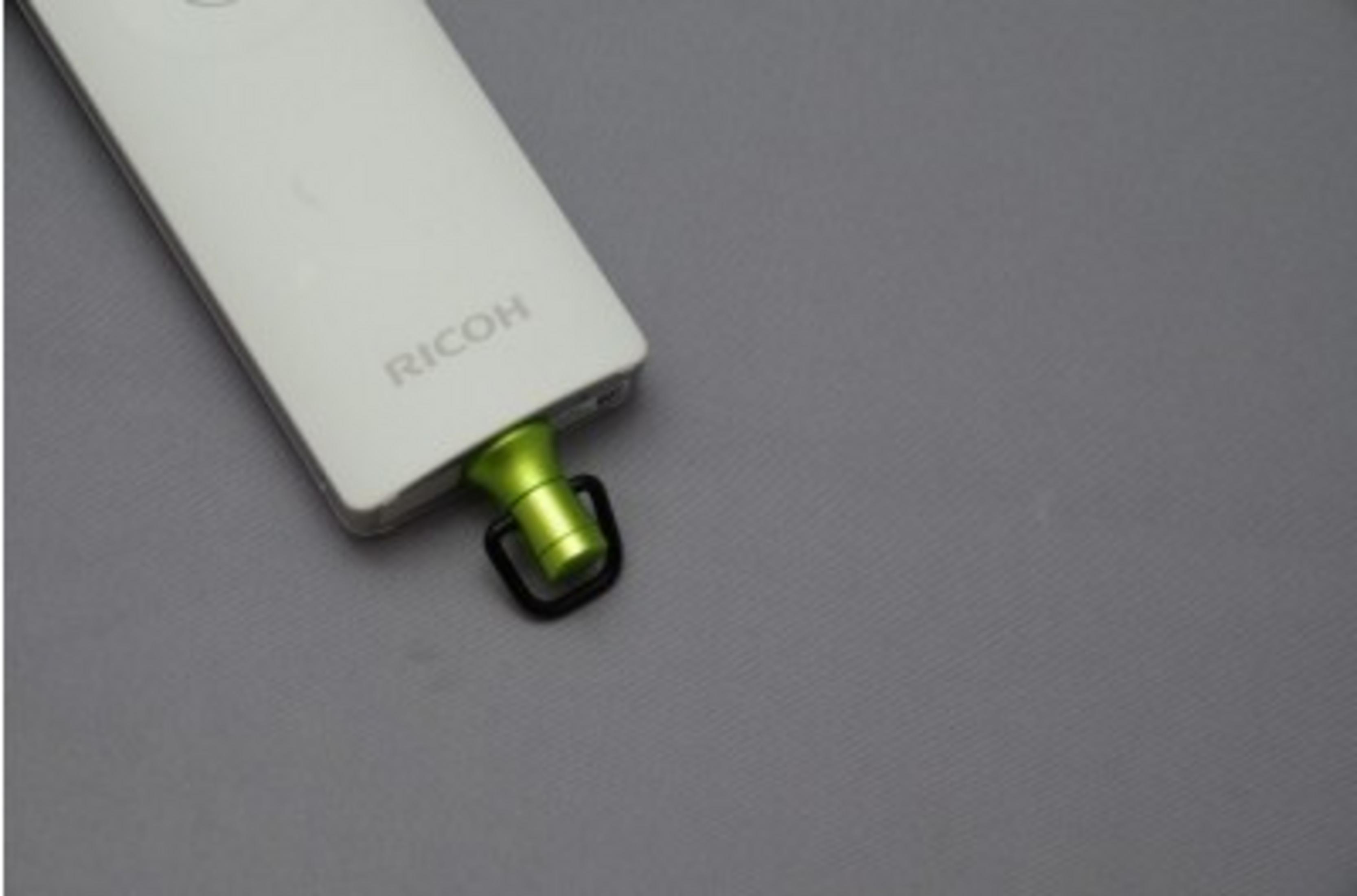 Białe urządzenie Ricoh z zielonym złączem i czarnym pierścieniem na szarym tle.