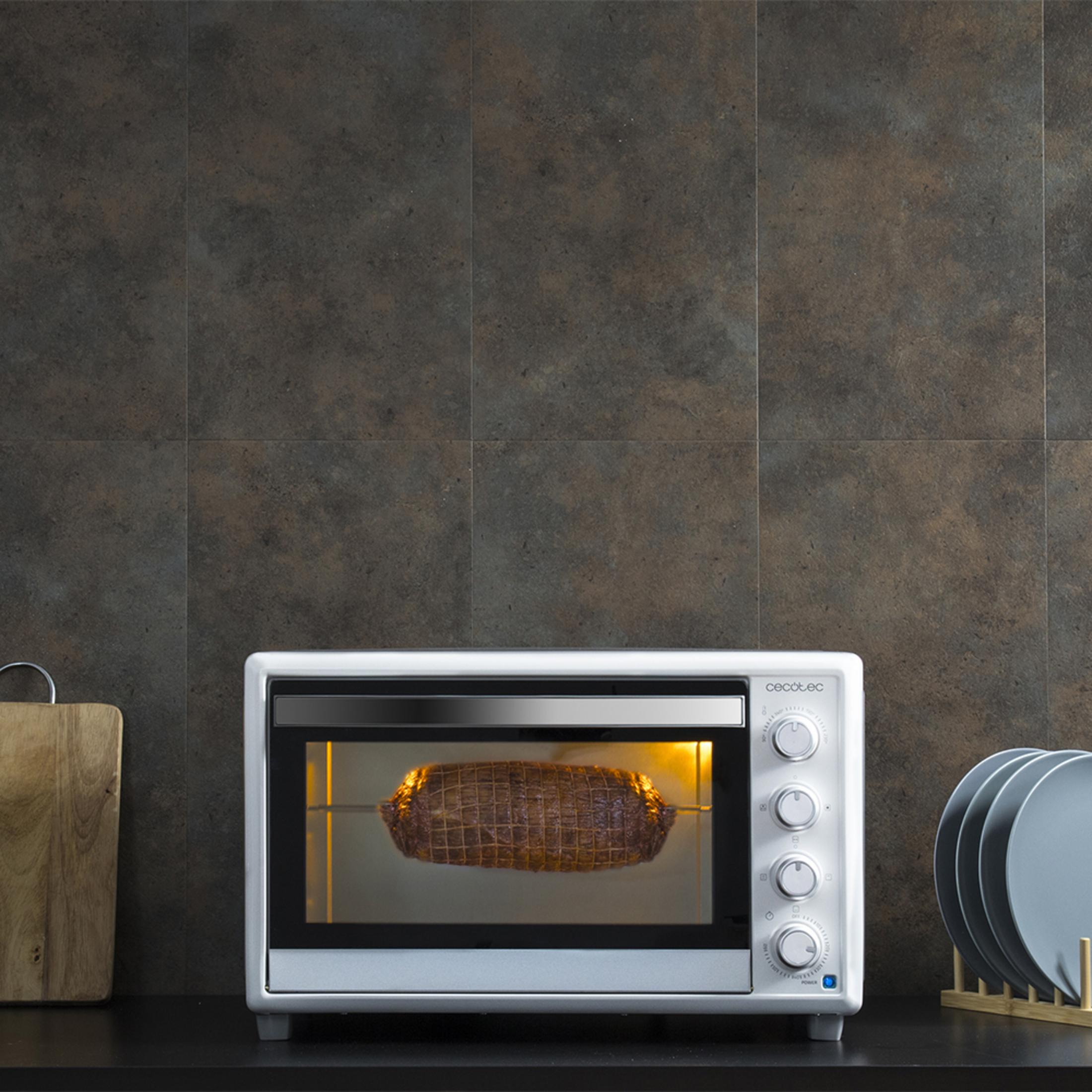 Un forno bianco arrostisce carne in rete. Vicino ci sono un tagliere e piatti.