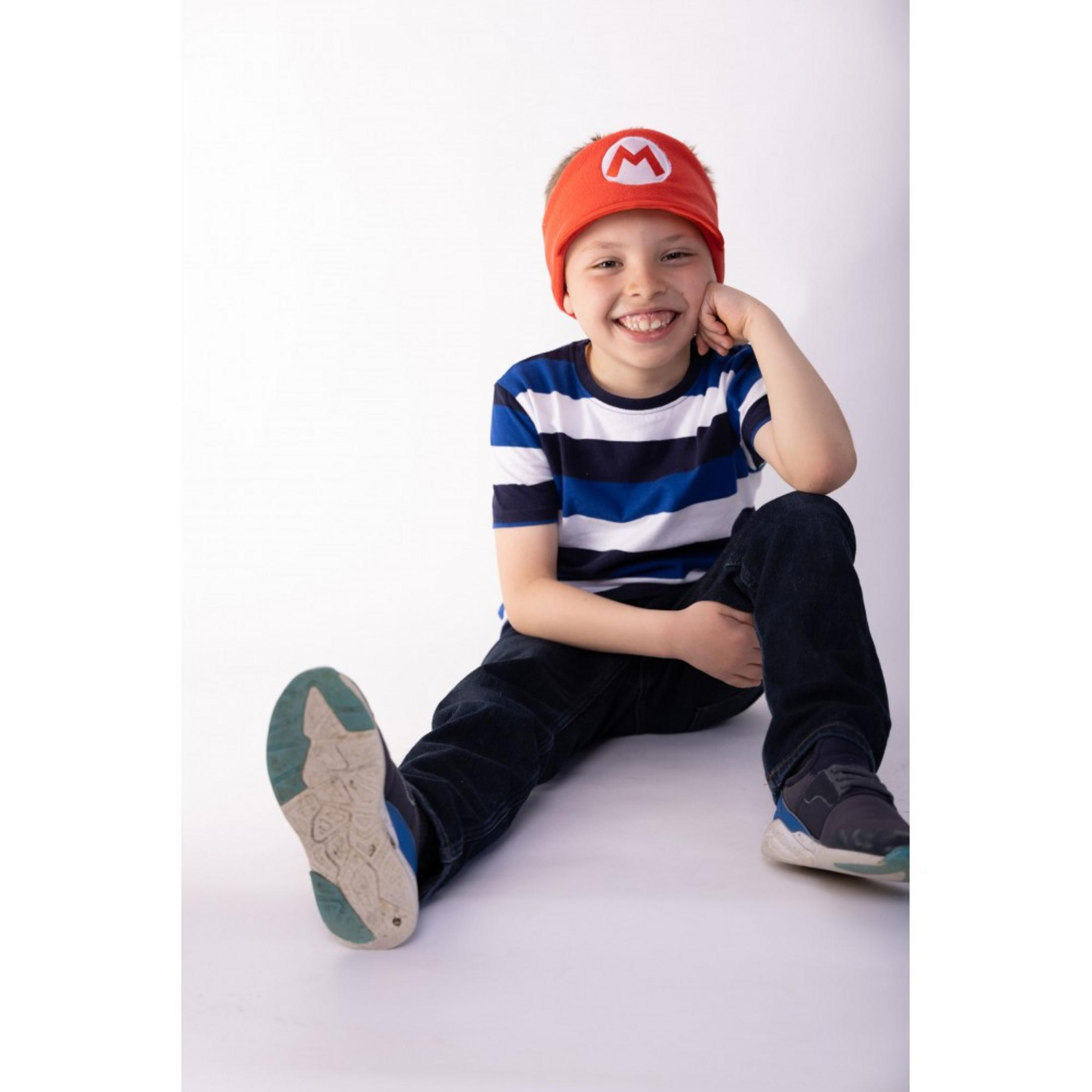 Un garçon assis par terre porte un bandeau Mario, un t-shirt rayé et un jean.