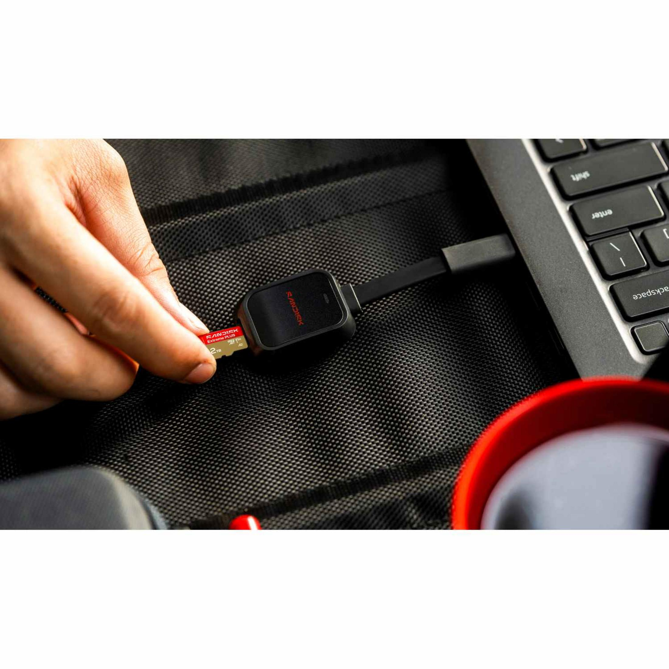 Main insère une carte SD dans un lecteur à côté d'un clavier et d'une tasse rouge.