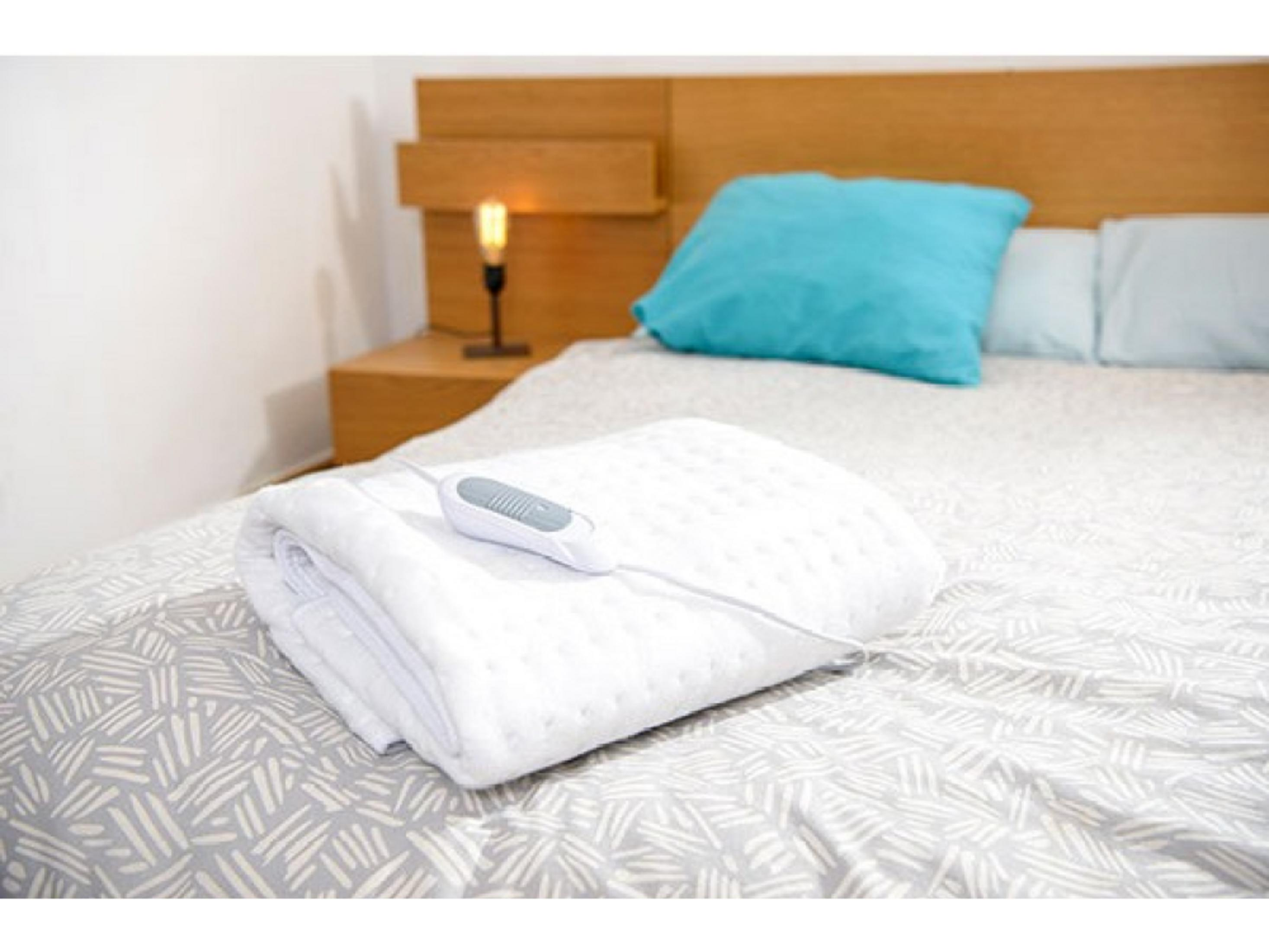 A white electric blanket with a controller lies on a bed.
