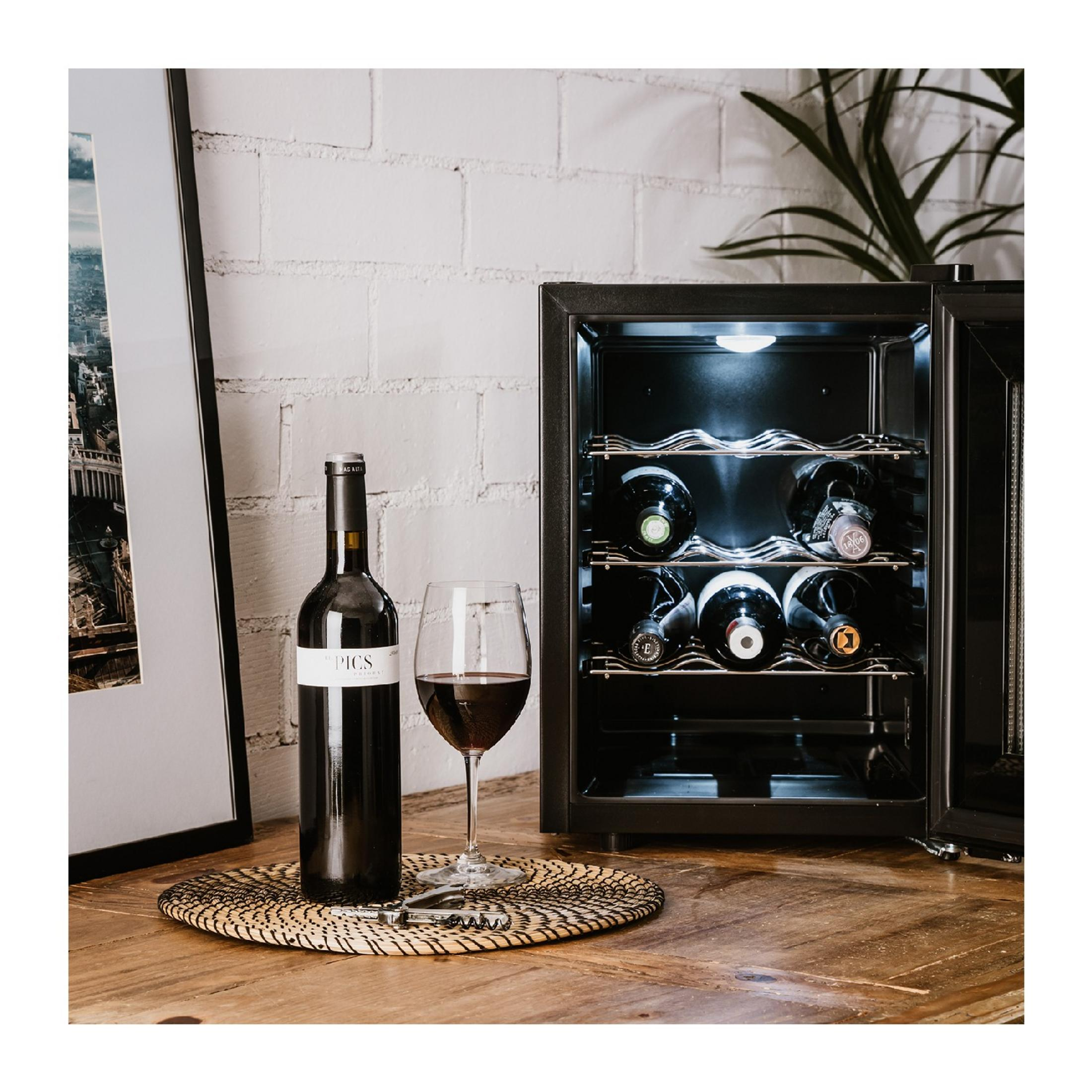 Una botella de vino y una copa de vino tinto junto a una vinoteca abierta sobre una mesa.