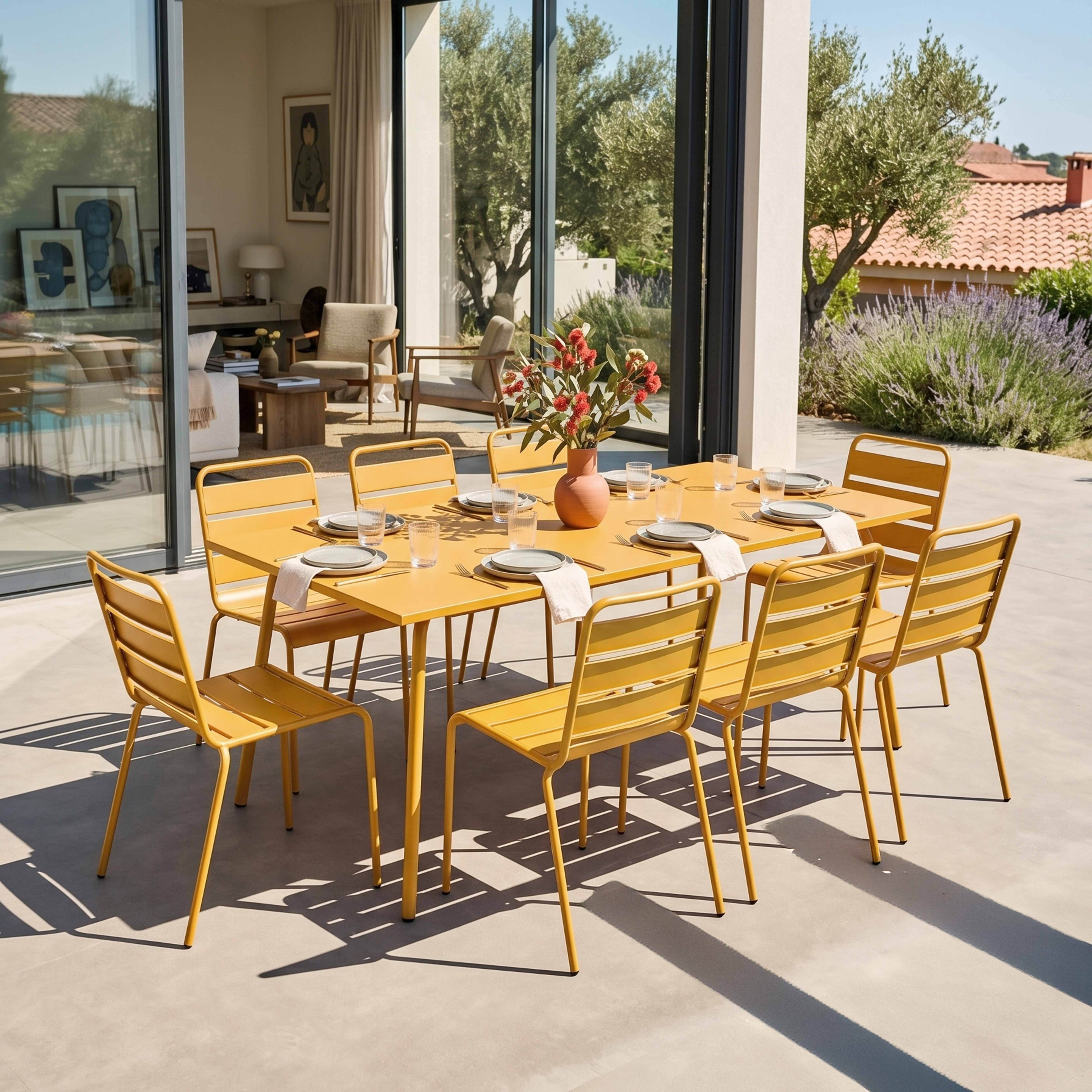 Ein gelber Esstisch mit Stühlen auf einer Terrasse, mit Blumen und Haus im Hintergrund.