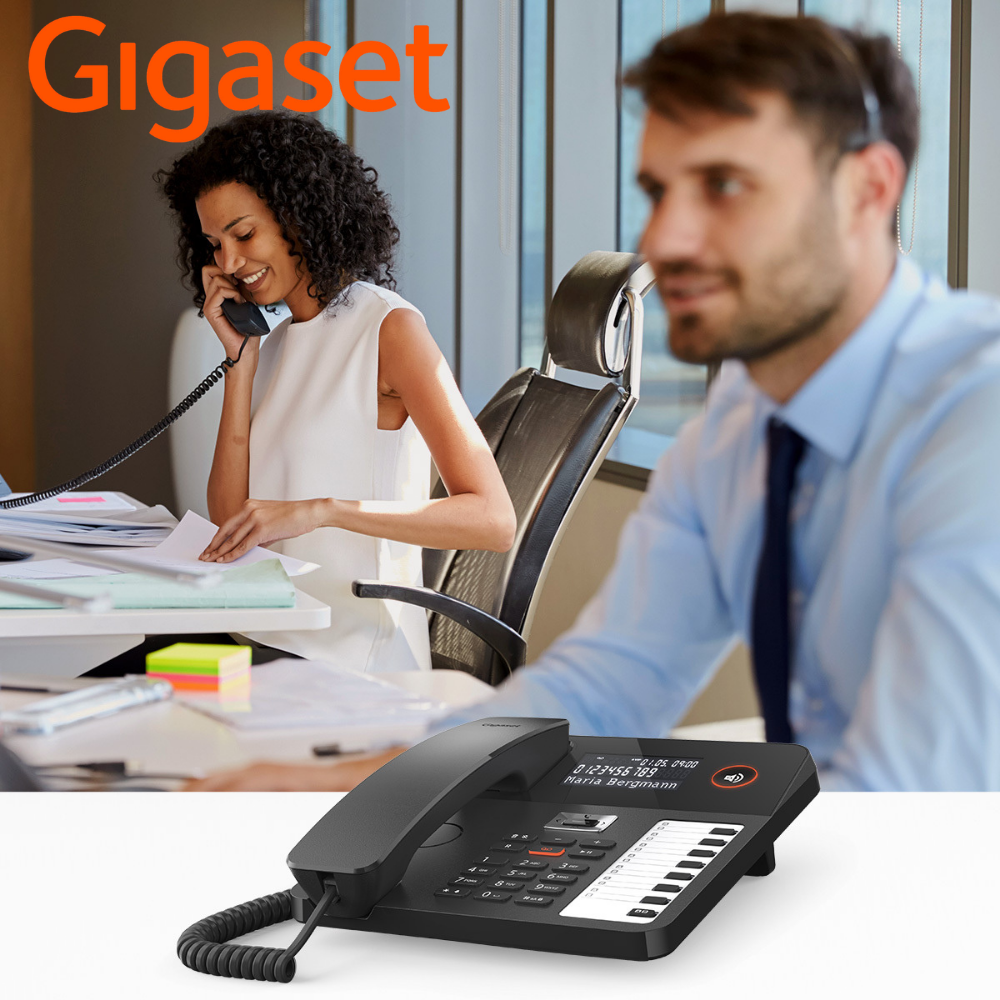 Ein Bürotelefon steht im Vordergrund, im Hintergrund telefonieren zwei Personen.