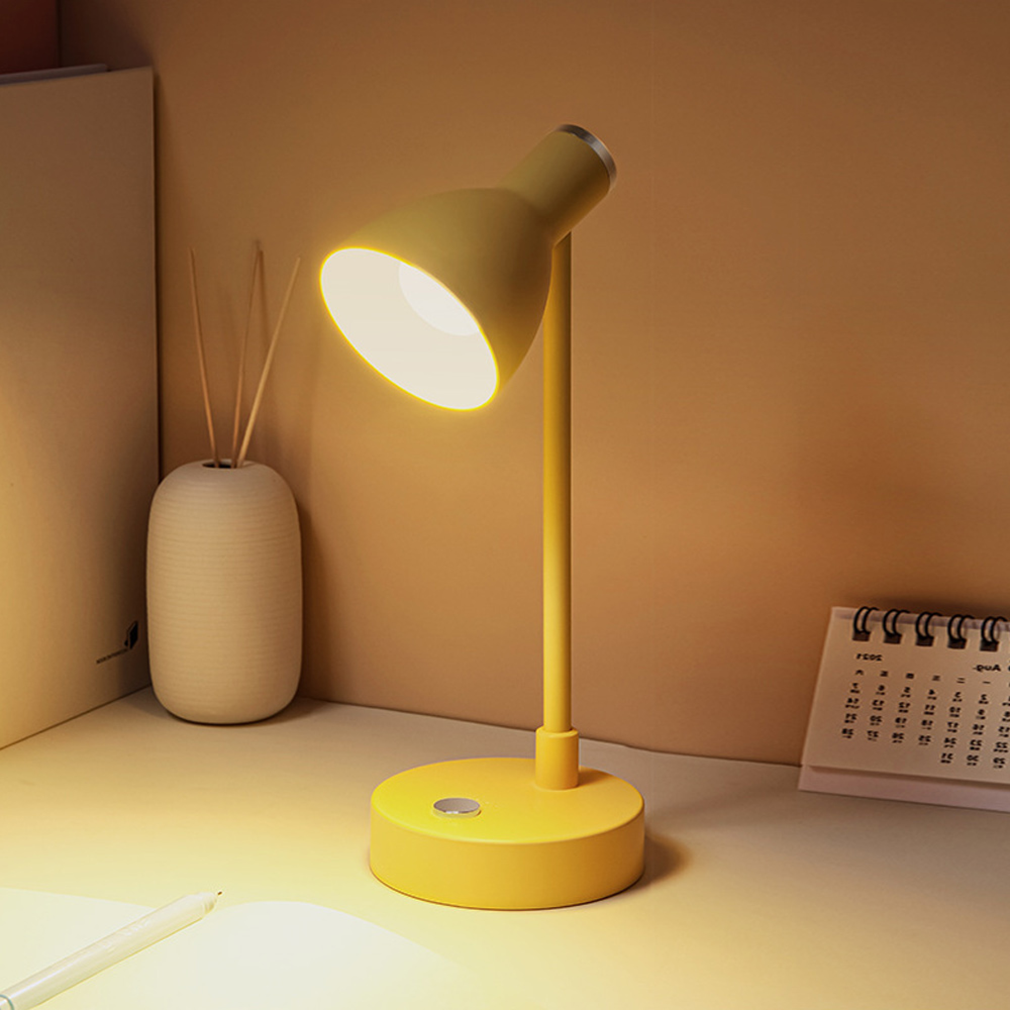 Eine gelbe Schreibtischlampe leuchtet auf einem Schreibtisch neben einem weißen Vasen-Diffusor.