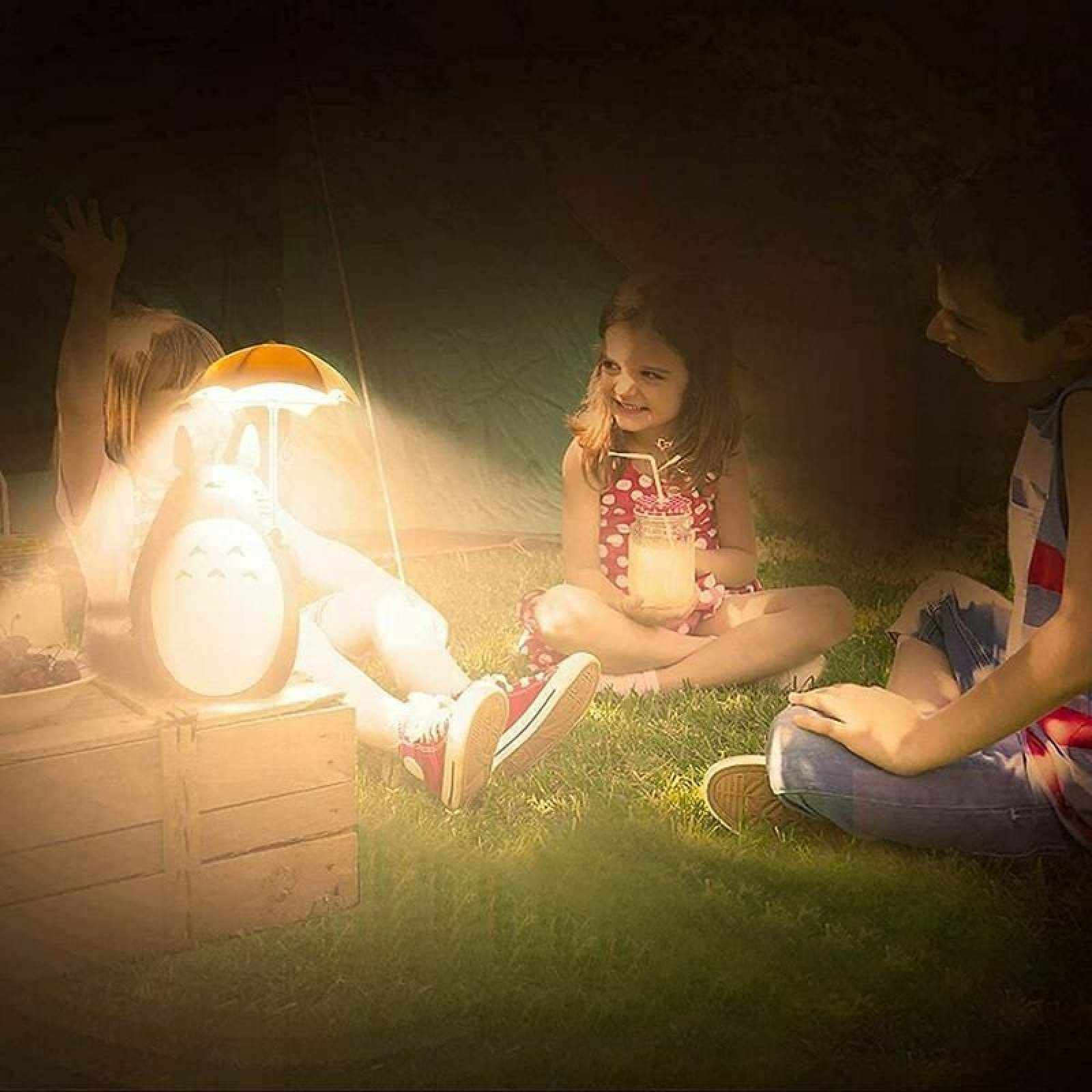 Drei Kinder sitzen auf Gras um eine Totoro-Lampe. Eines hält ein Getränk.