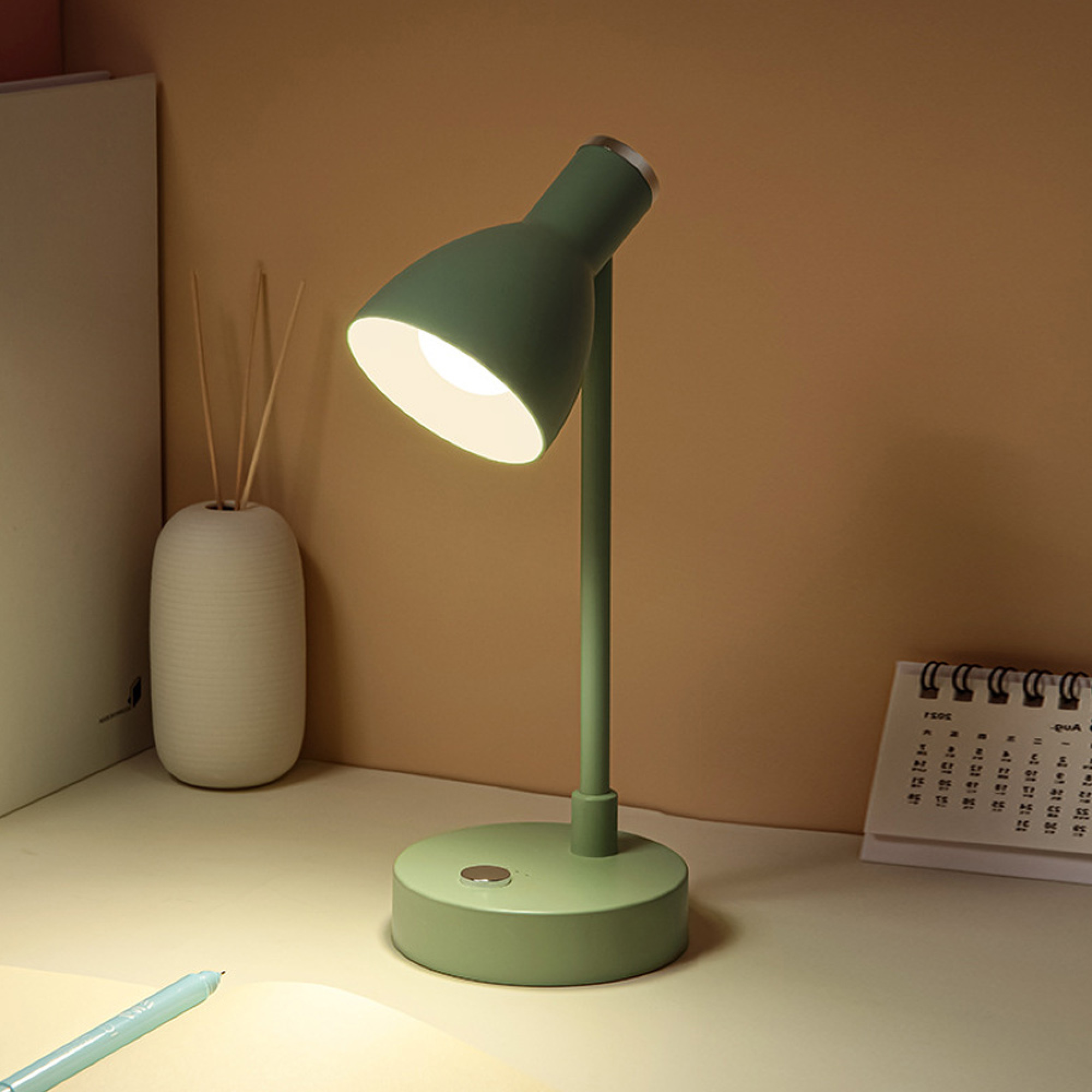 Grüne Schreibtischlampe leuchtet auf einem Schreibtisch neben einem Kalender und einer Vase.