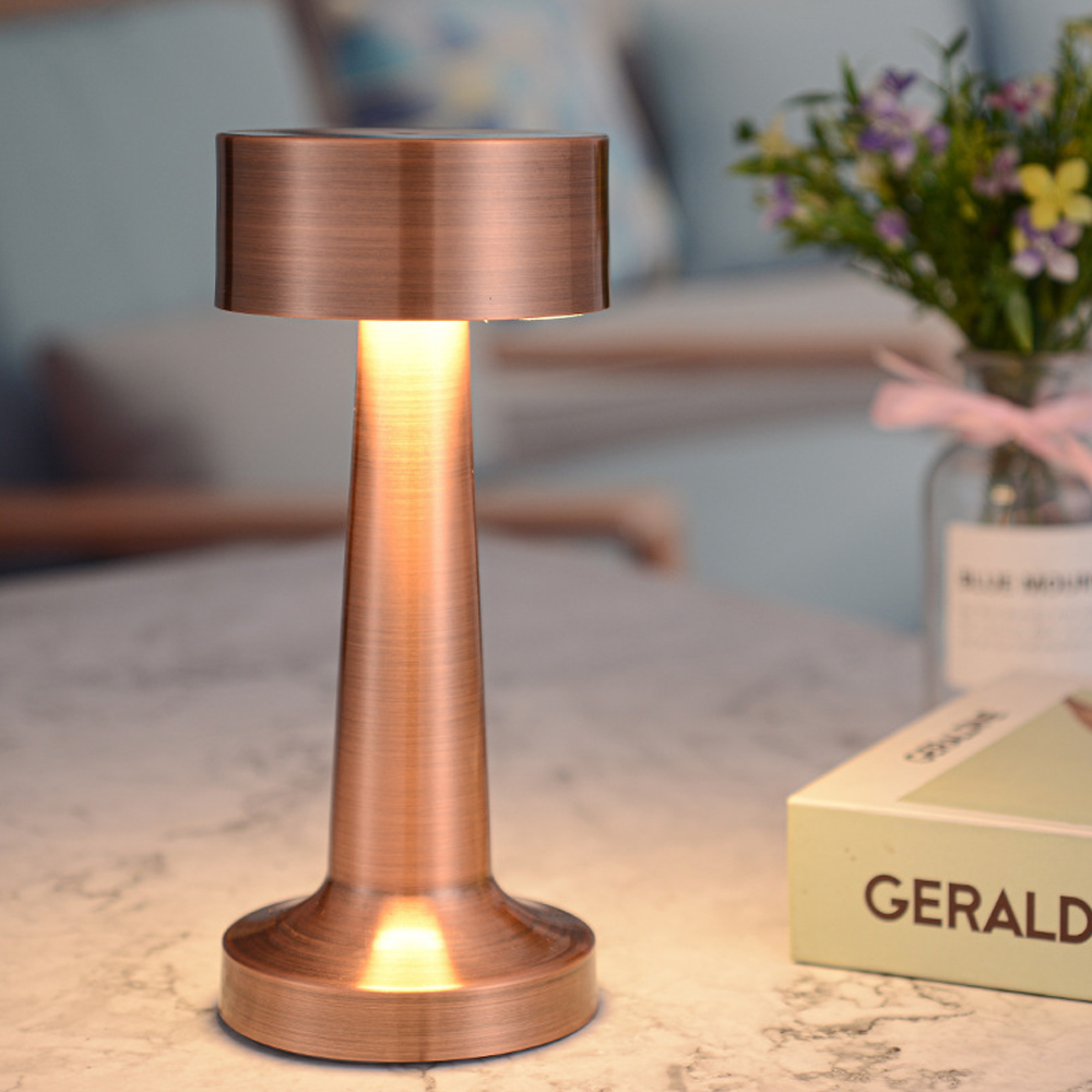 Eine bronzene Tischlampe leuchtet auf einem Marmortisch, daneben ein Buch und Blumen.