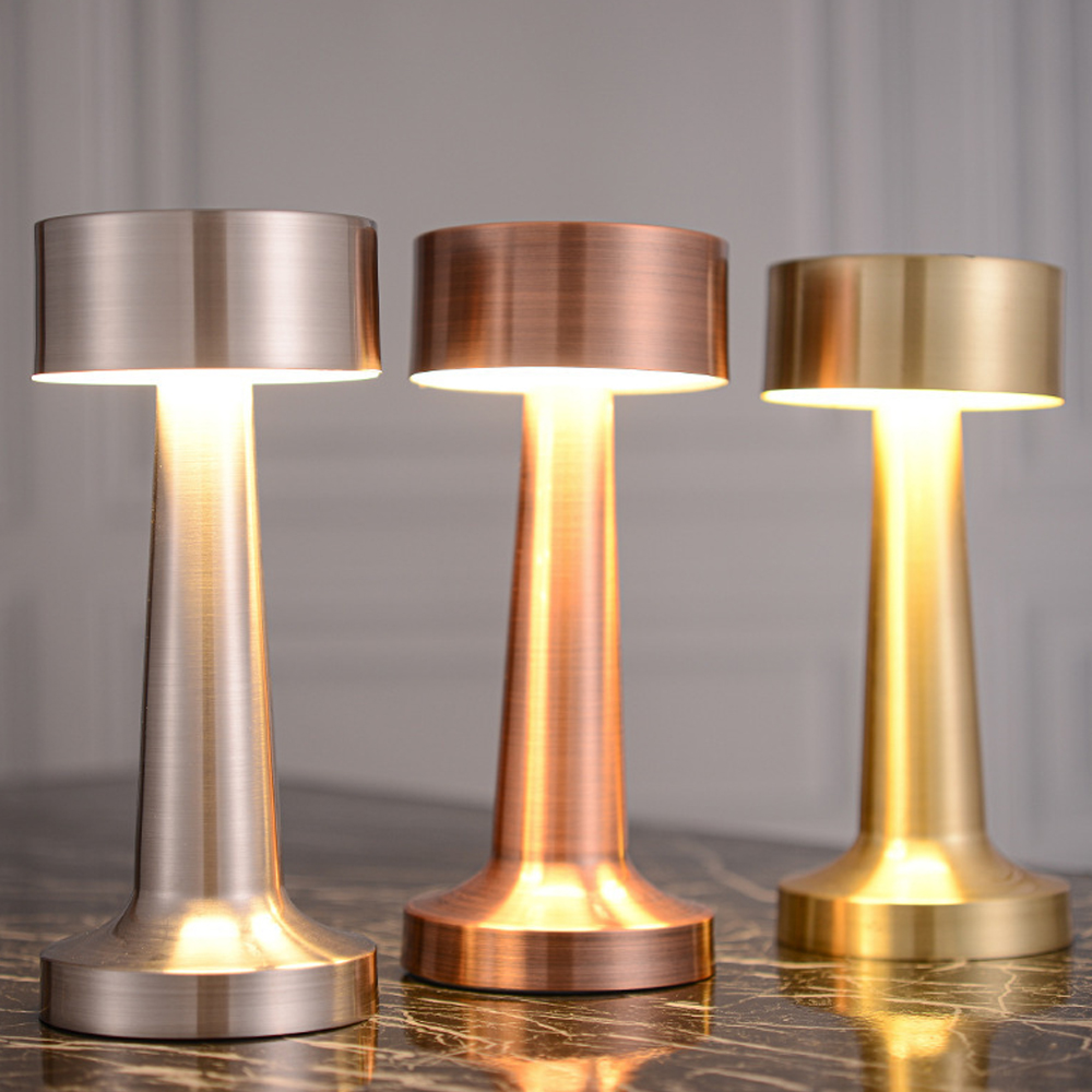 Drei moderne Tischlampen in Silber, Kupfer und Gold stehen nebeneinander.