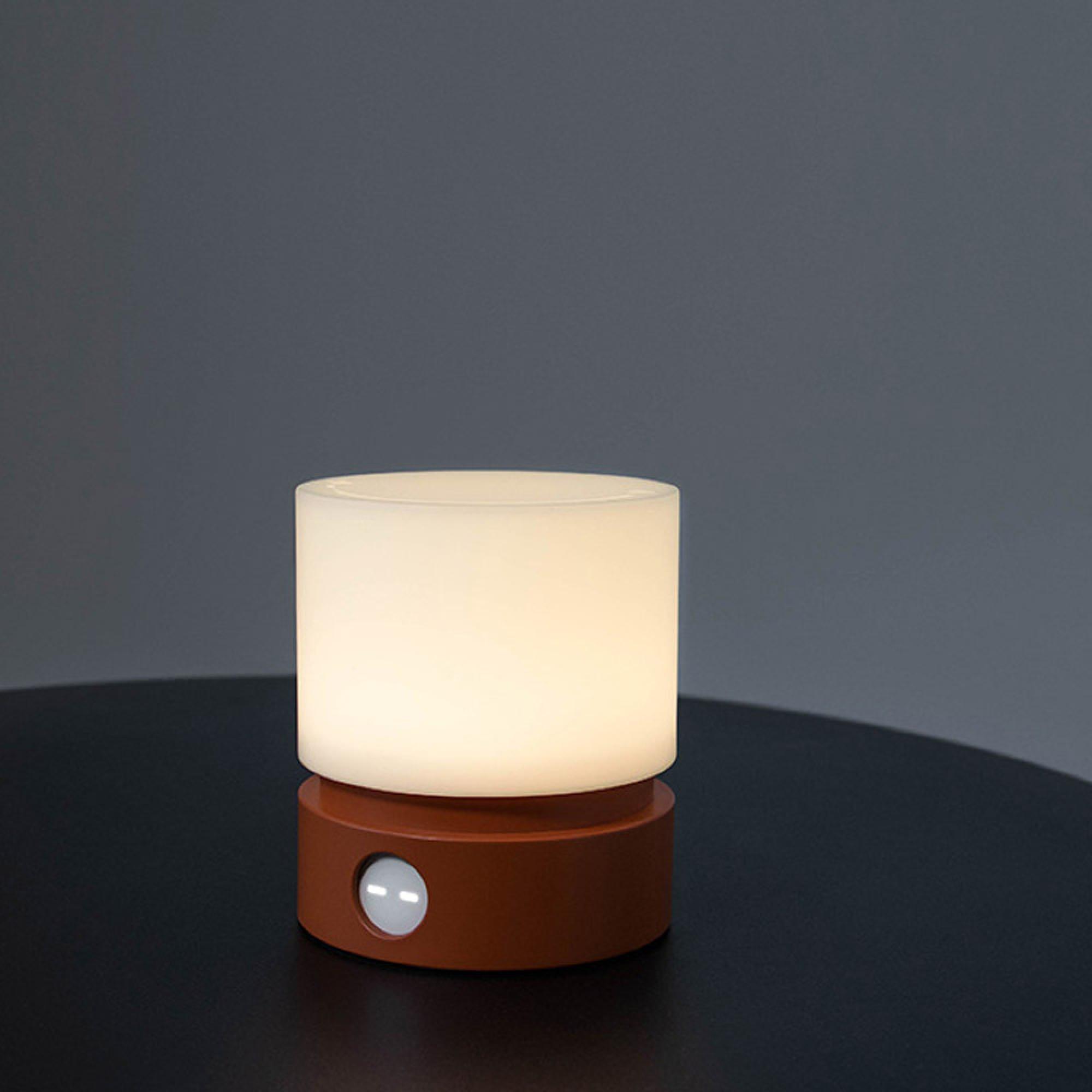 Eine leuchtende weiße Lampe mit braunem Sockel und Sensor auf dunklem Tisch.