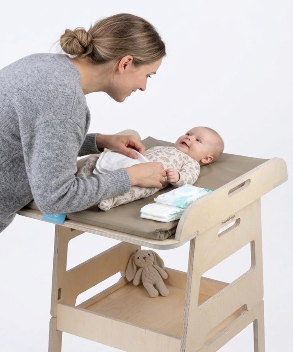Anne, ahşap bebek bakım masasında bebeğinin bezini değiştiriyor. Rafta peluş tavşan var.