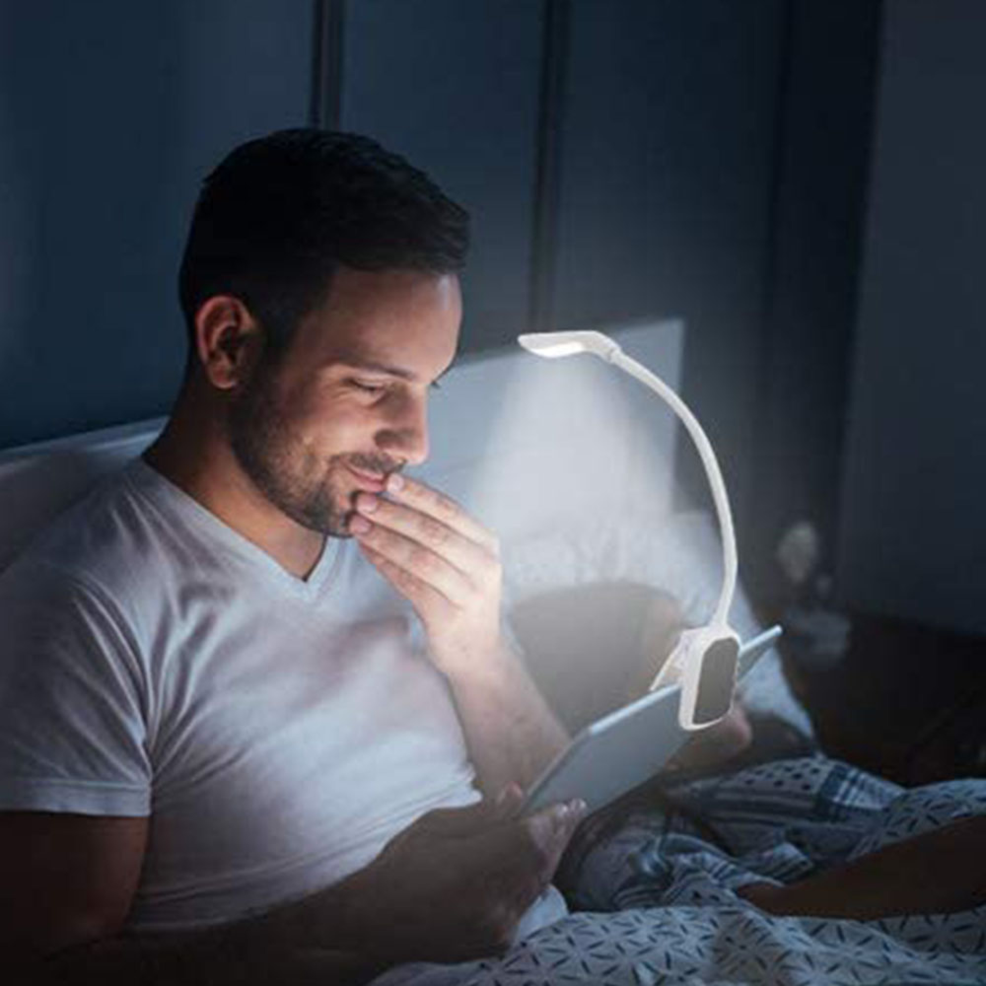 Ein Mann liegt im Bett und liest auf einem Tablet, beleuchtet von einer Klemmlampe.