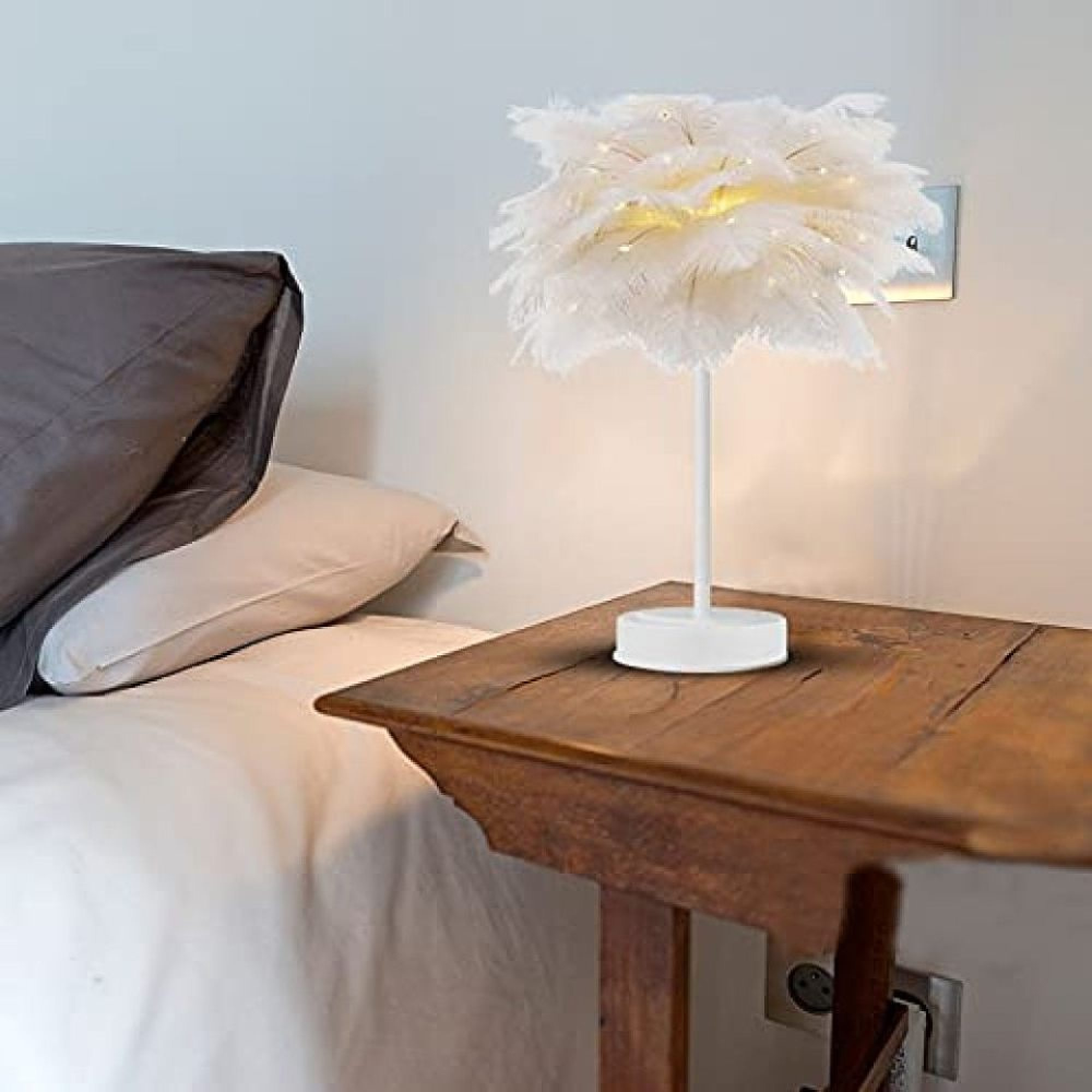 Eine weiße Federlampe mit Lichtern steht auf einem Nachttisch neben einem Bett.