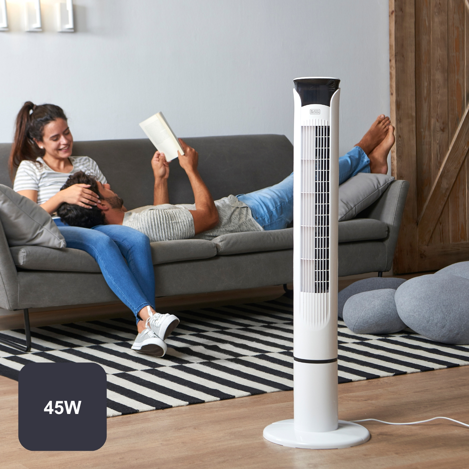 Una pareja relajándose en un sofá con un ventilador de torre cerca.