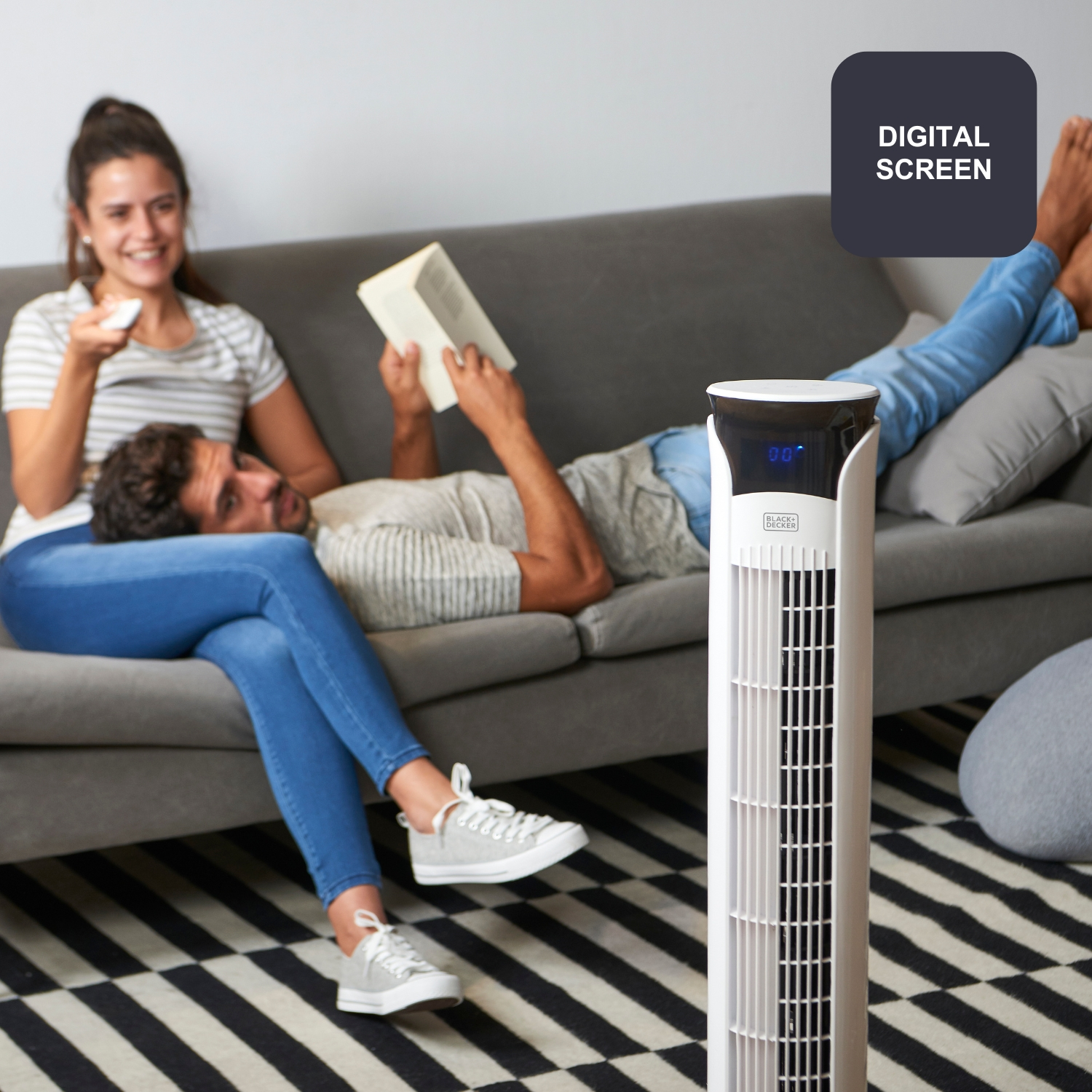 Una pareja relajándose en un sofá con un ventilador de torre cerca.