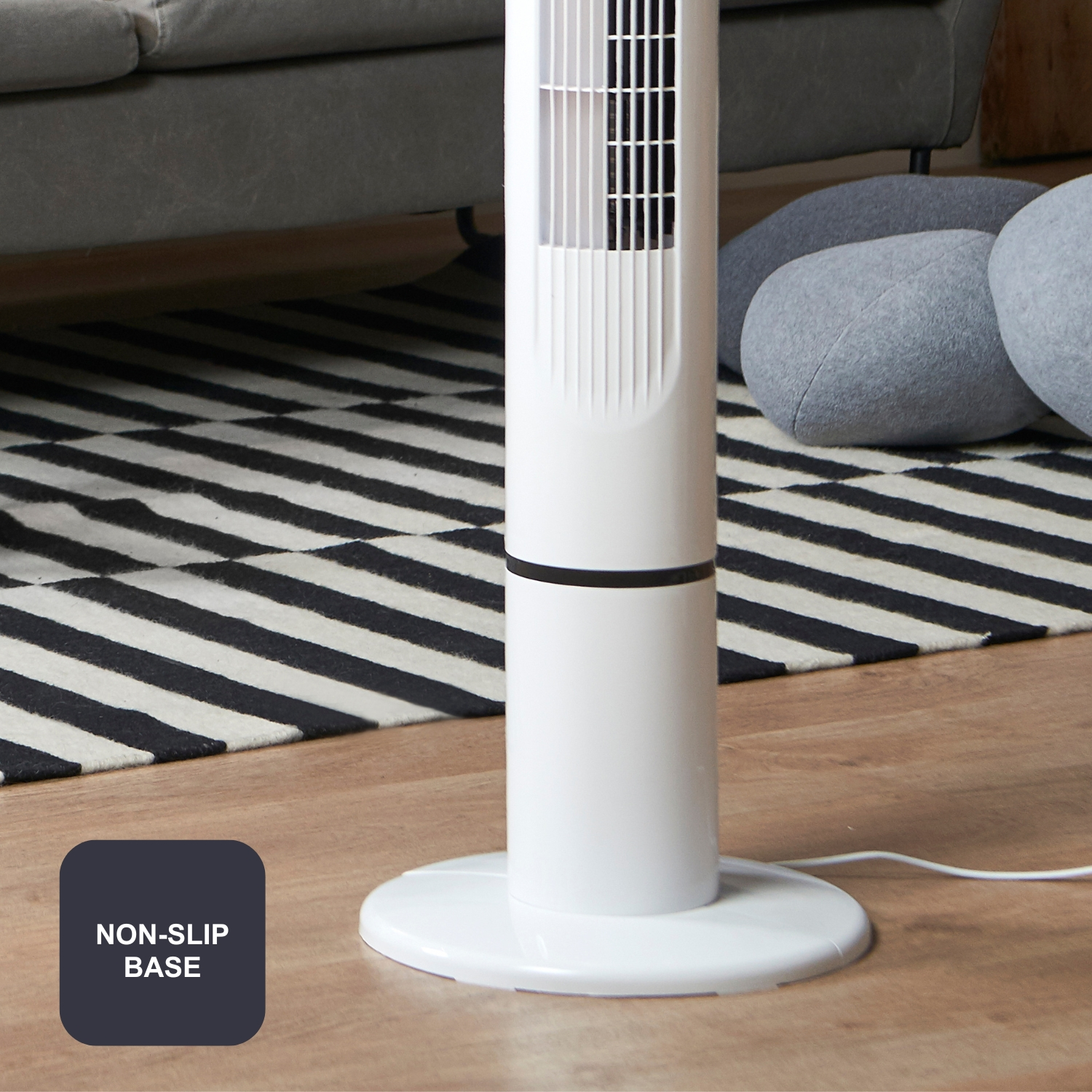 Un ventilador de torre blanco con base antideslizante sobre suelo de madera junto a alfombra.