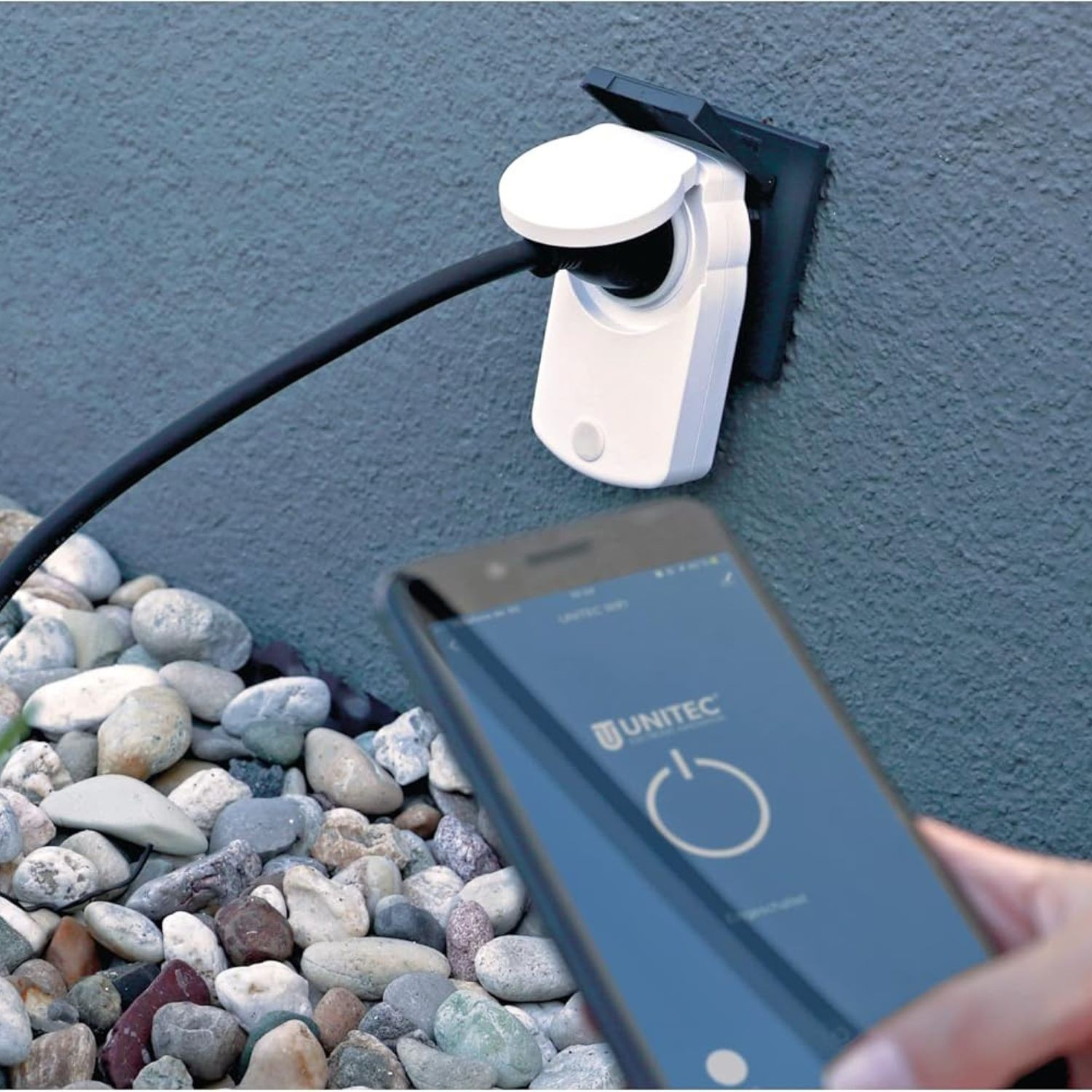 Eine Hand hält ein Telefon mit UNITEC-App. Ein Smart-Stecker an der Wand ist mit Kabel verbunden.