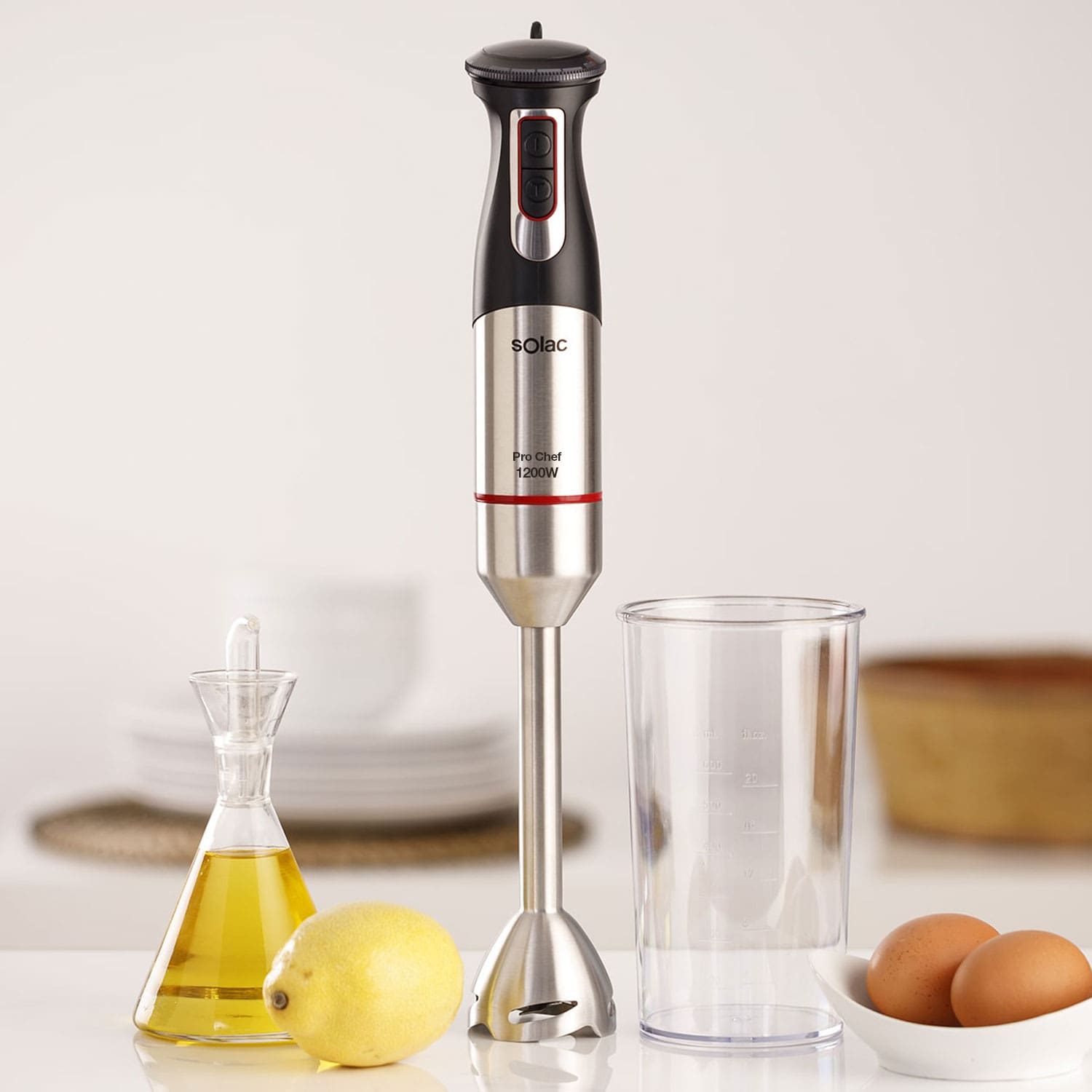 Una batidora de inmersión con accesorios: aceite, limón, huevos, vaso medidor en una mesa.