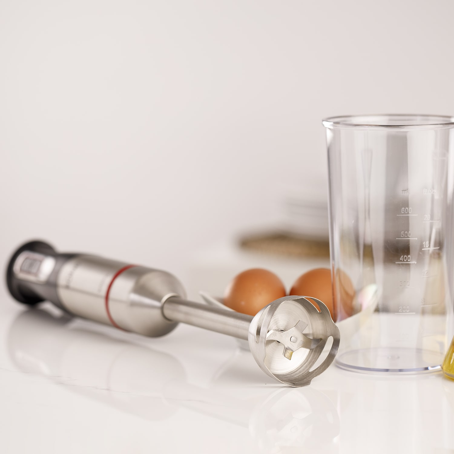 Una batidora de inmersión descansa junto a huevos y un vaso medidor en una encimera.