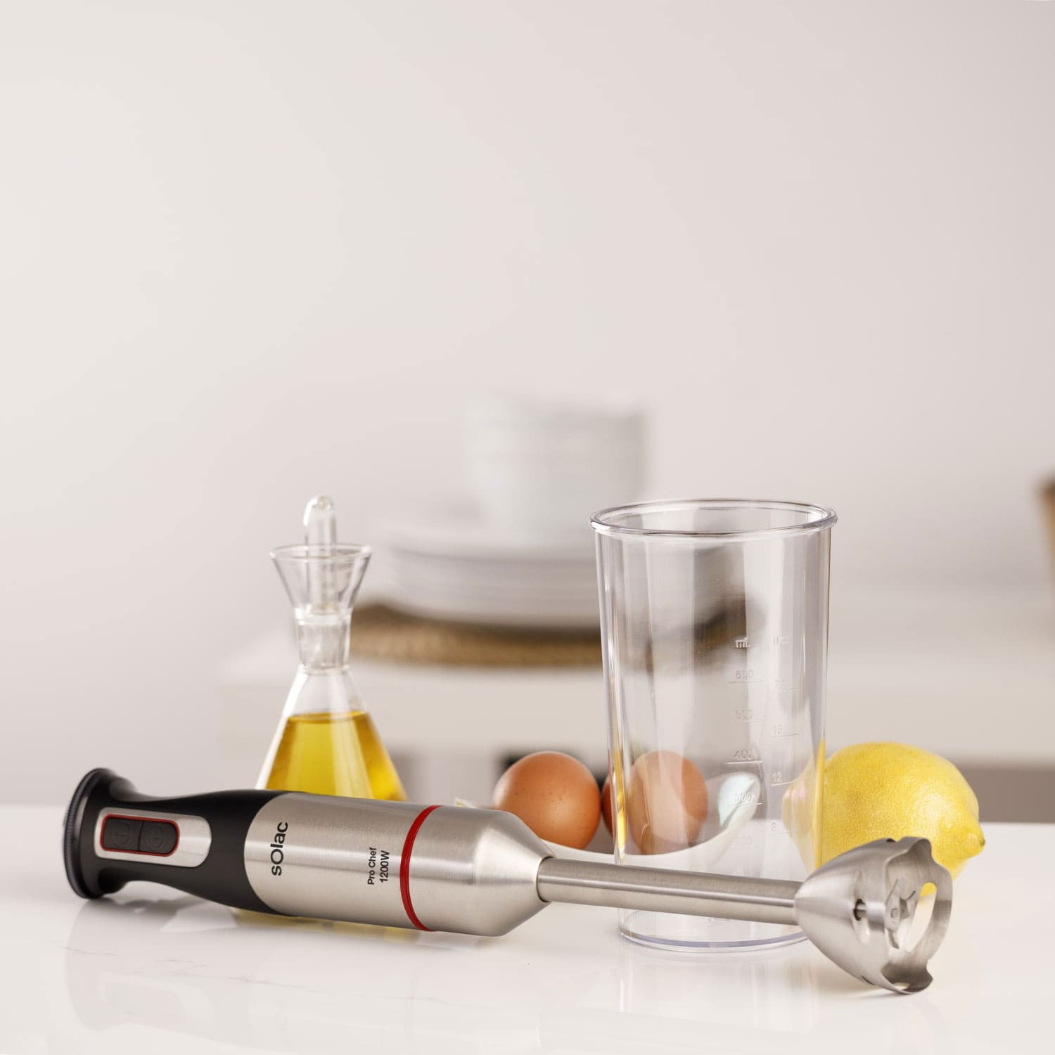 Una batidora de mano yace sobre una superficie blanca junto a huevos, limón, aceite y un vaso medidor.