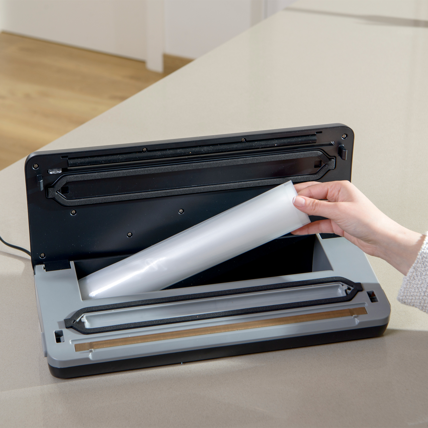Una mano coloca un rollo de material de bolsa de plástico transparente en una selladora al vacío gris y negra.