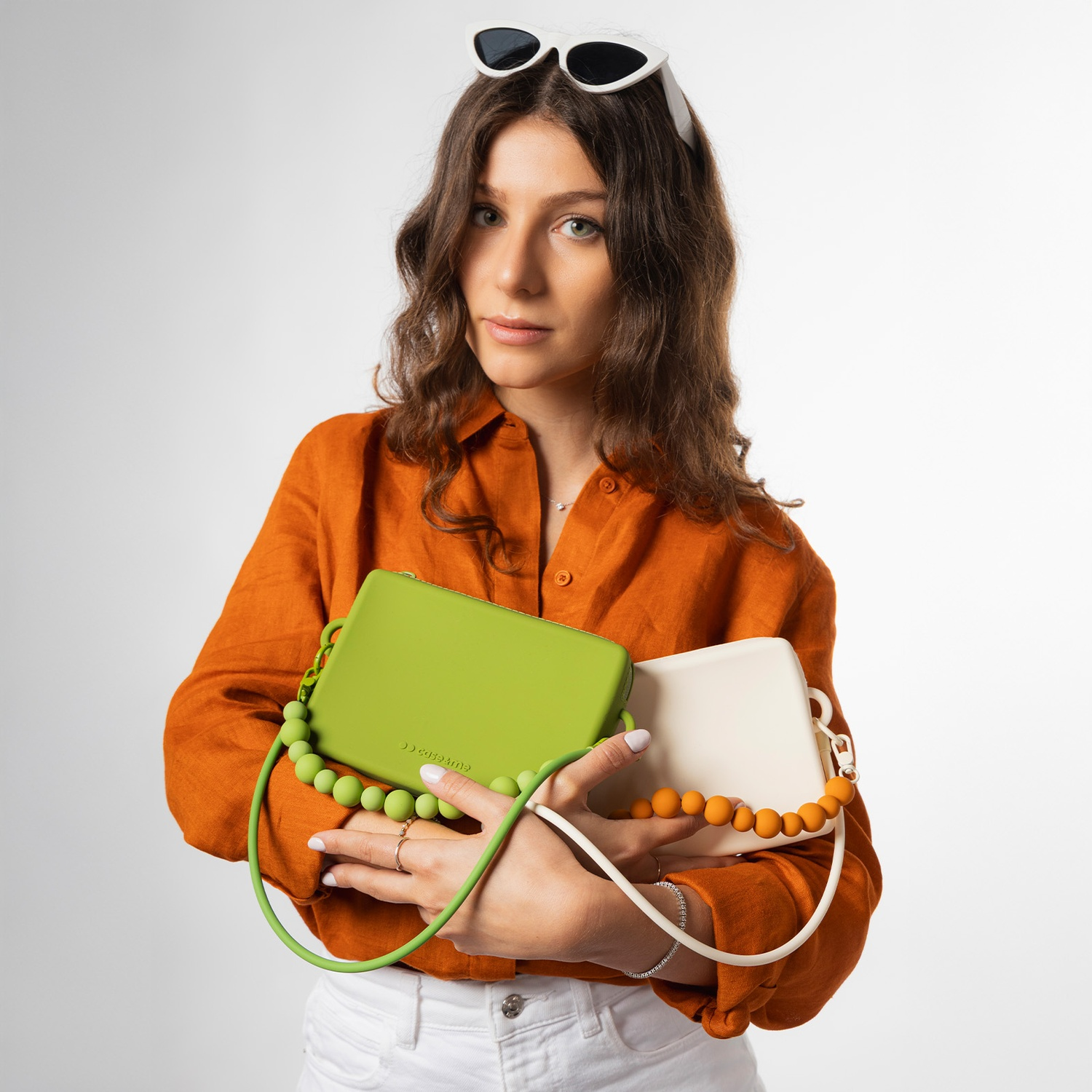 Una mujer con cabello castaño y gafas de sol blancas sostiene un bolso verde y uno color crema.