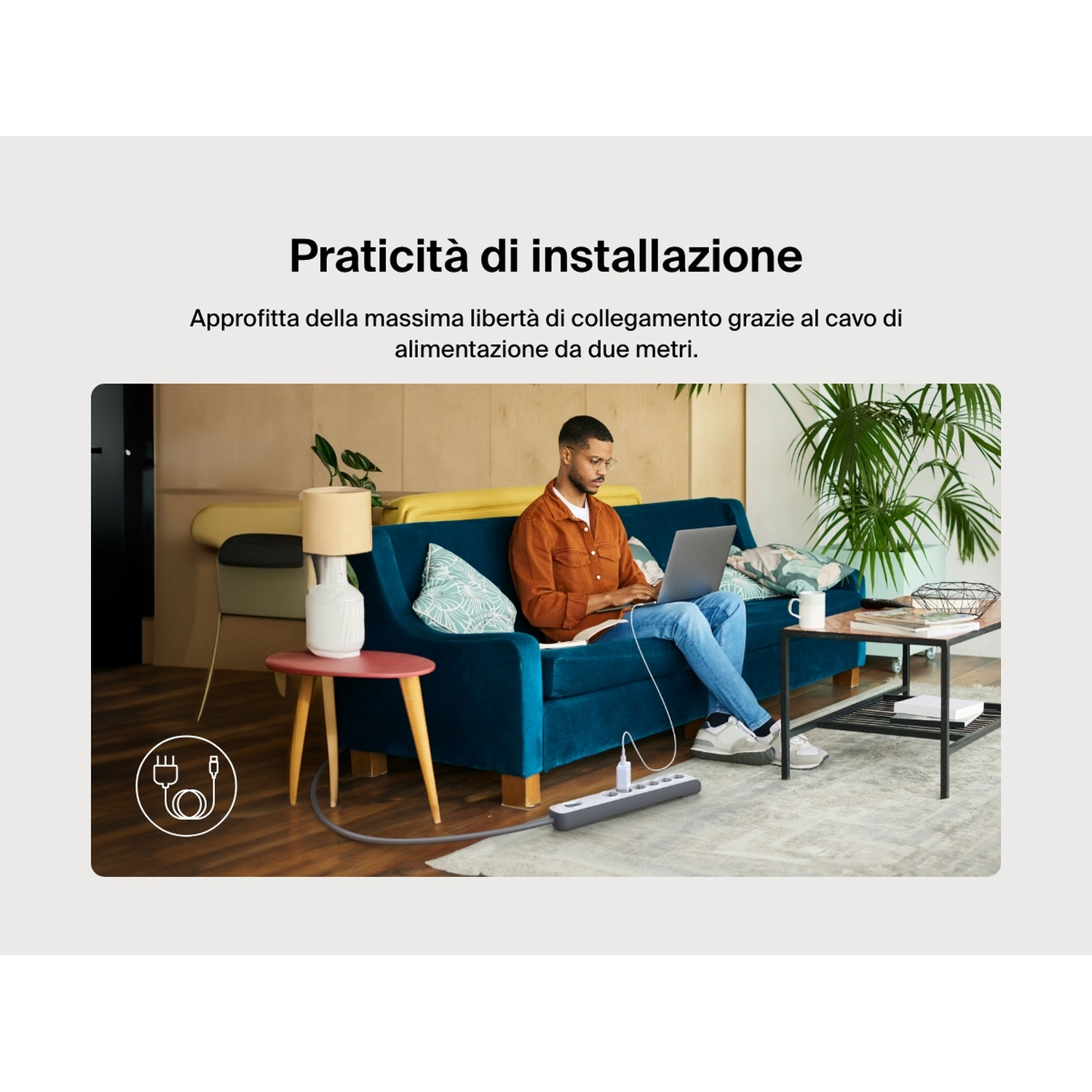 Uomo sul divano con laptop, ciabatta elettrica e lampada.