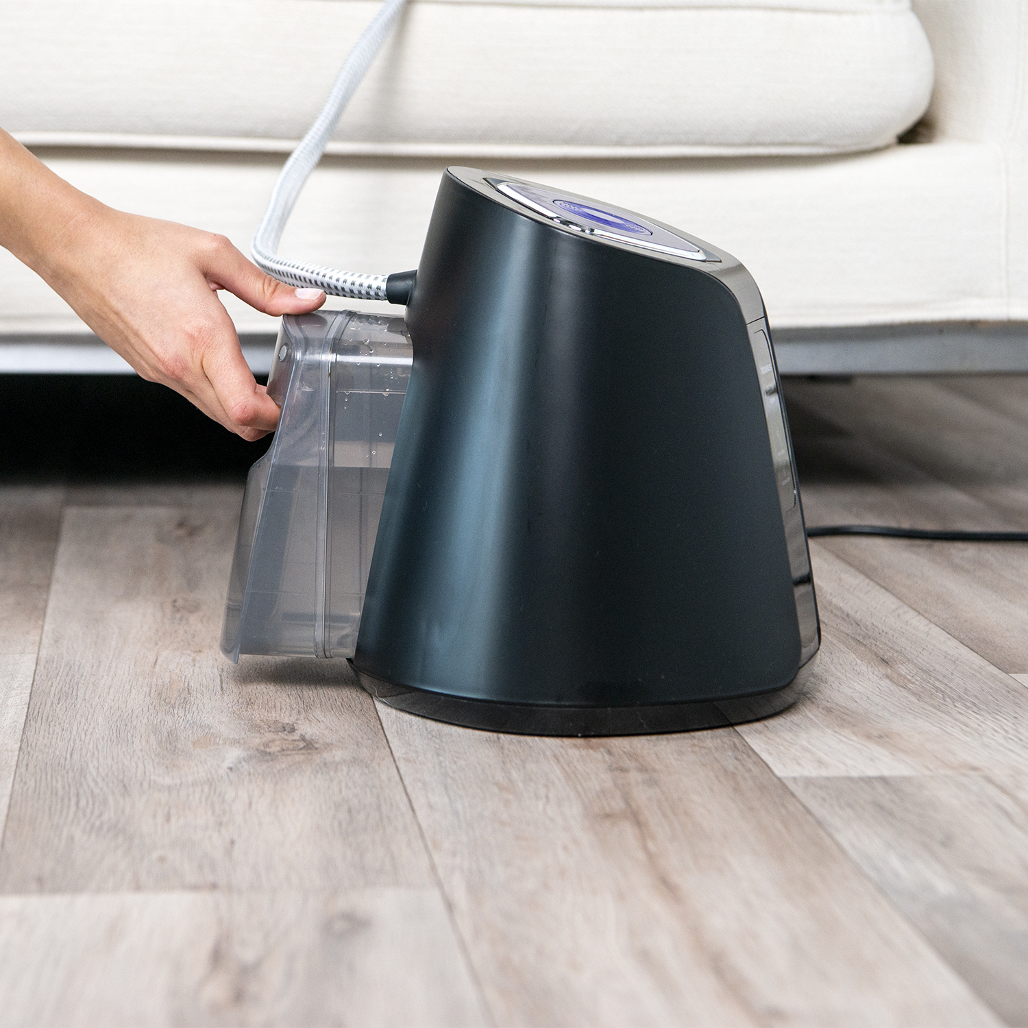 Una mano coloca un depósito de agua transparente en un limpiador a vapor negro sobre suelo de madera.