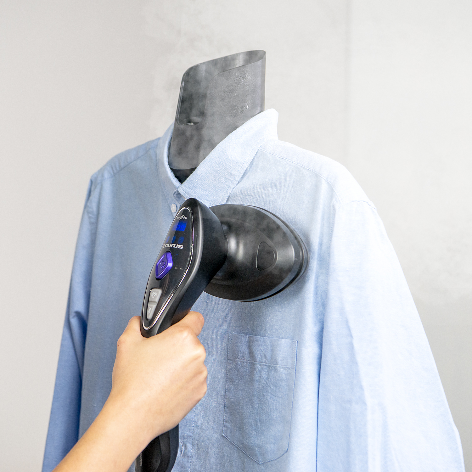 Una mano sostiene un vaporizador de ropa, alisando una camisa azul en un maniquí.