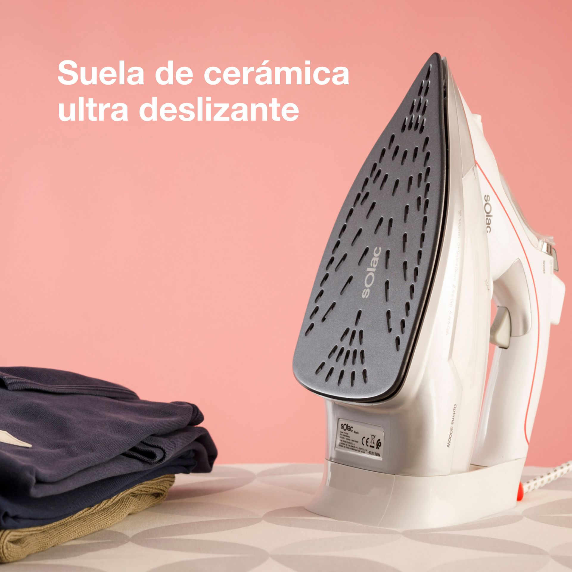 Una plancha blanca con suela de cerámica y una pila de ropa oscura sobre una mesa.