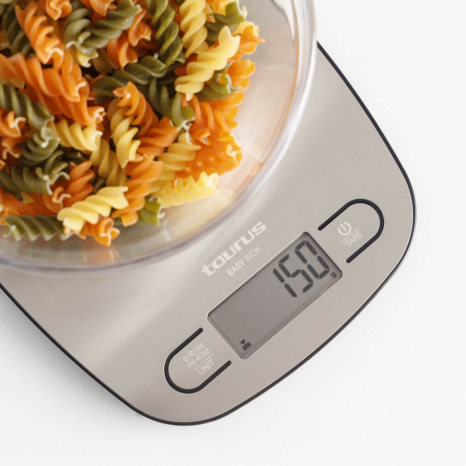 Un tazón de pasta rotini tricolor está sobre una báscula de cocina digital que marca 150 g.