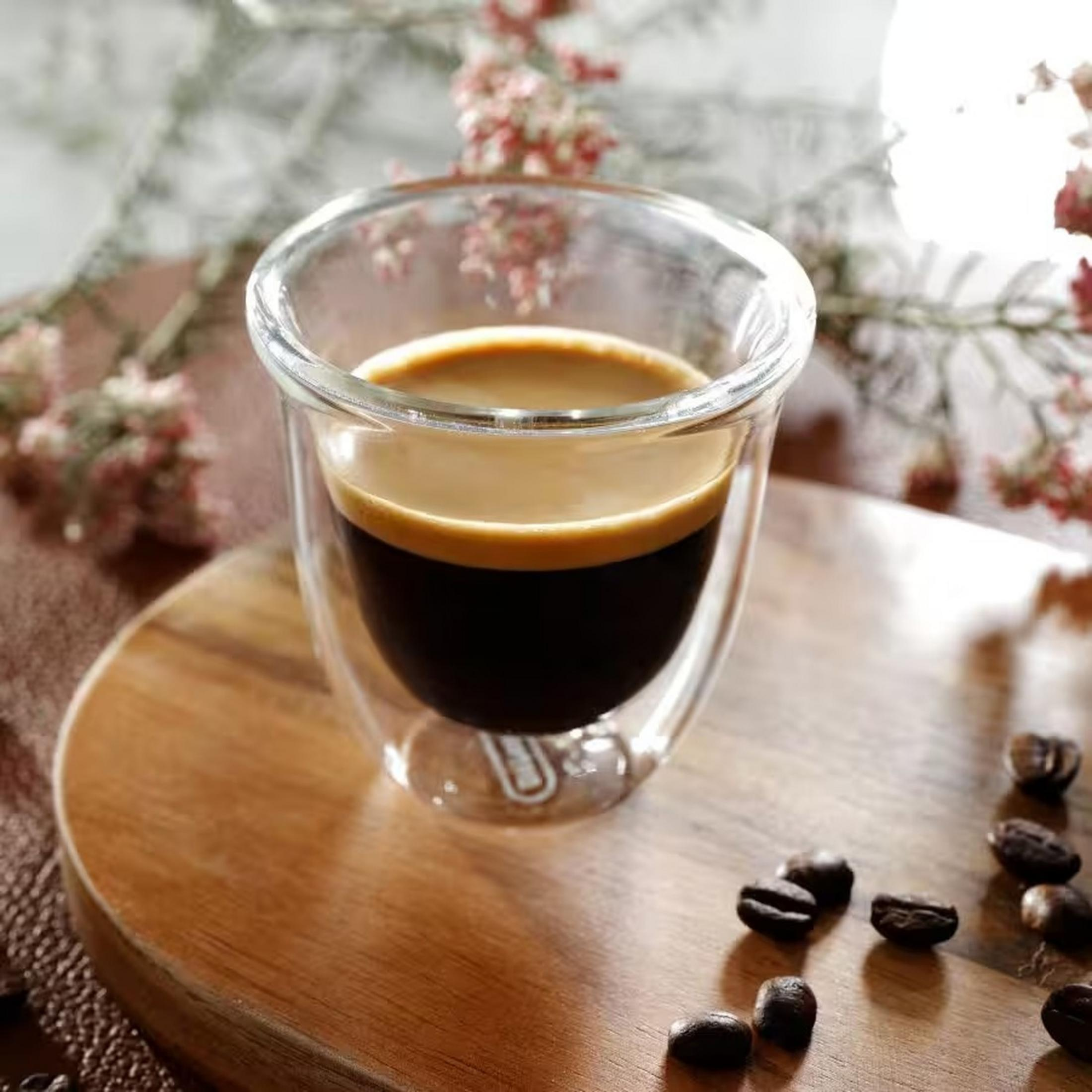 Ein doppelwandiges Glas hält dunklen Espresso mit cremiger Crema. Kaffeebohnen sind auf einem Holzbrett verstreut.