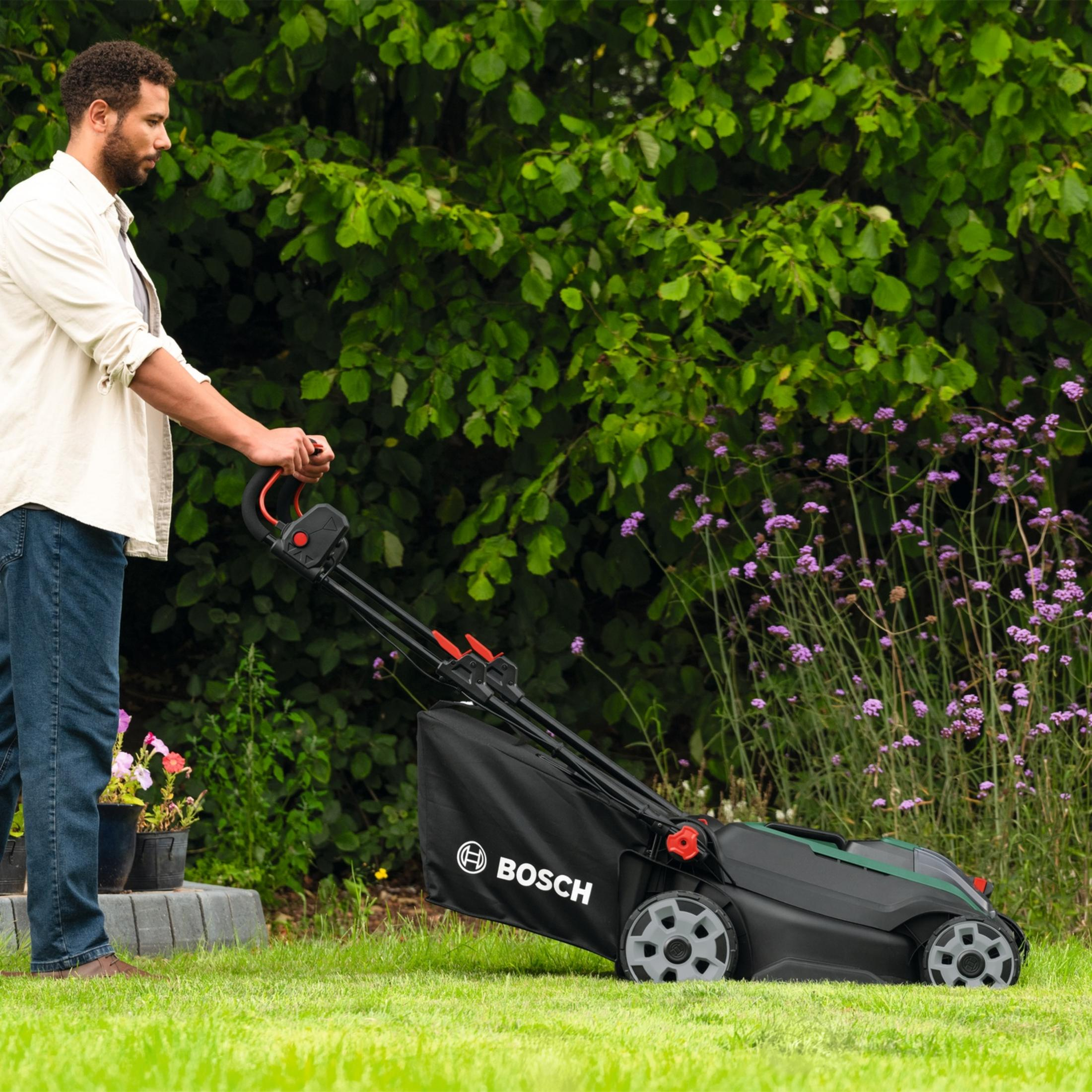 Mann schiebt einen Bosch-Rasenmäher auf einem grünen Rasen neben lila Blumen.