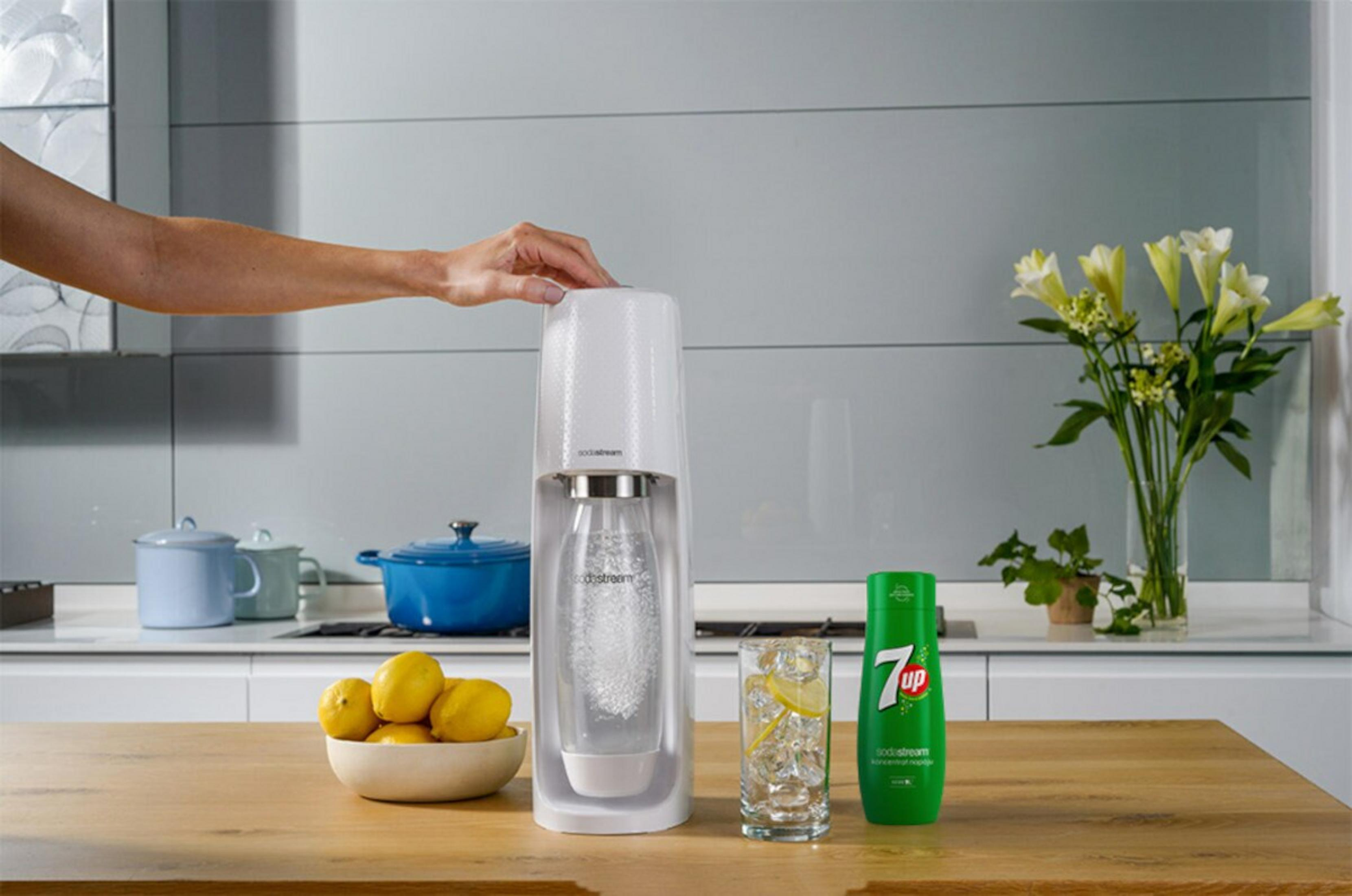 Eine Hand benutzt einen weißen Sodastream-Sprudler. Daneben ein Glas mit Eis und Zitrone, eine 7UP-Flasche, Zitronen und Blumen.