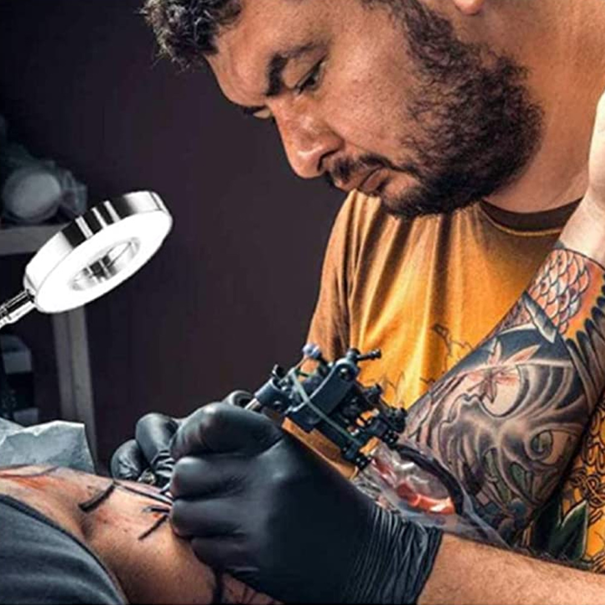 Ein Tätowierer mit schwarzer Handschuh-Hand benutzt eine Tätowiermaschine auf dem Unterarm eines Kunden.