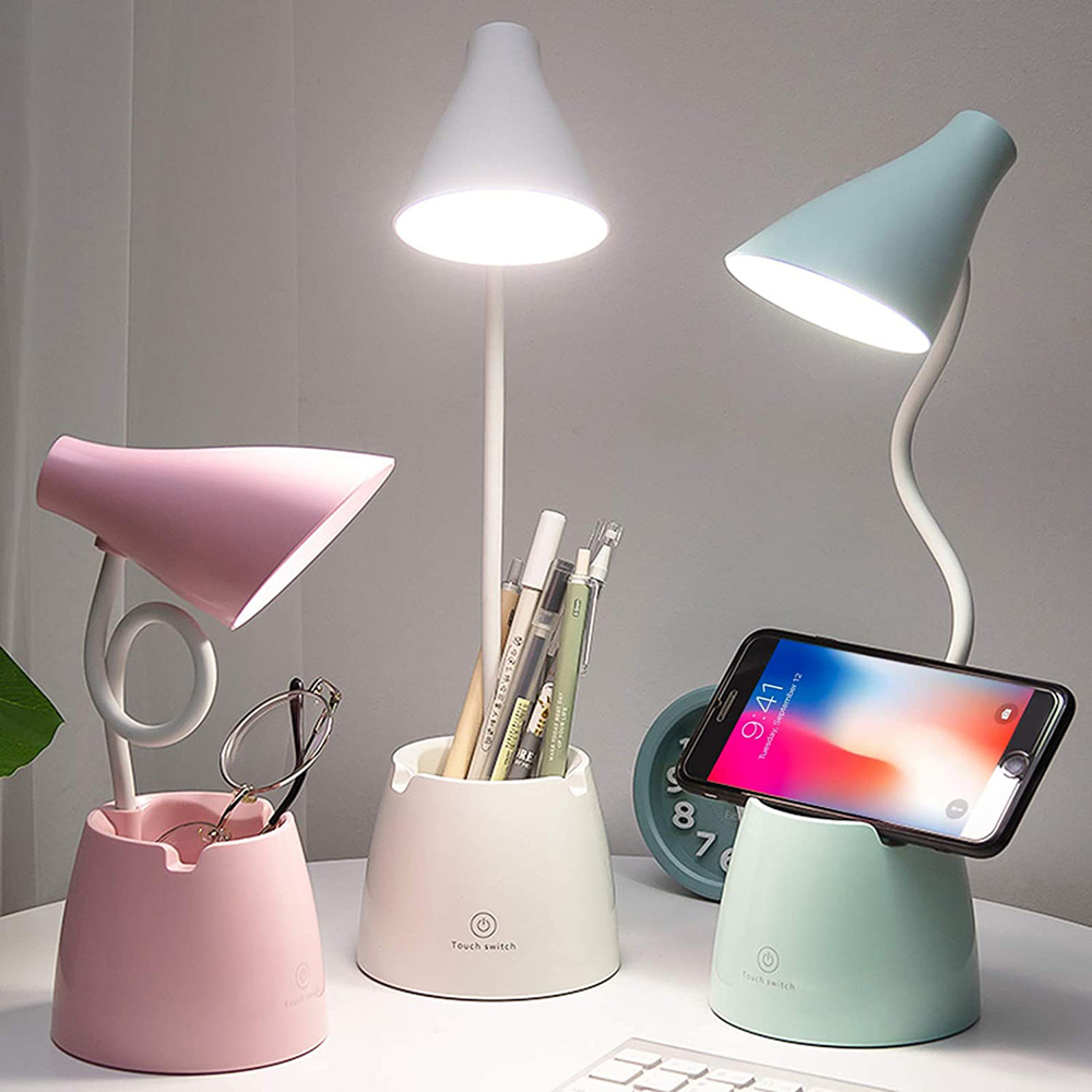 Drei Schreibtischlampen in Rosa, Beige und Hellgrün, mit Handy, Brille und Stiften.