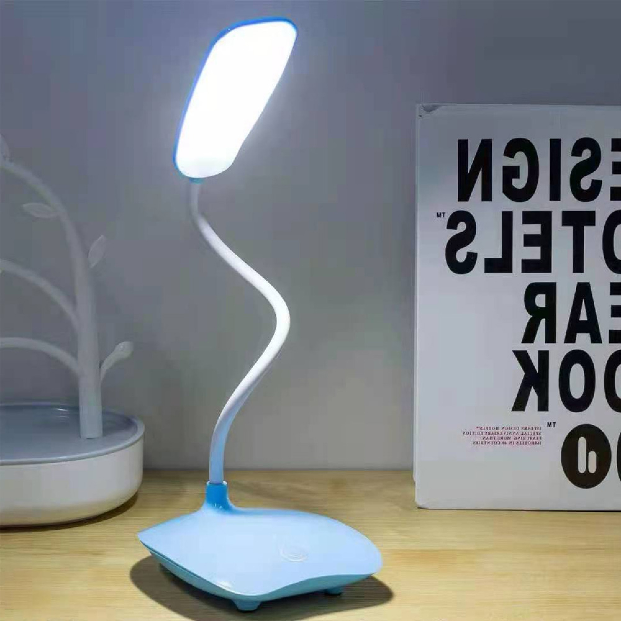 Eine blaue Schreibtischlampe mit biegsamem Arm steht auf einem Holztisch neben einem Buch.