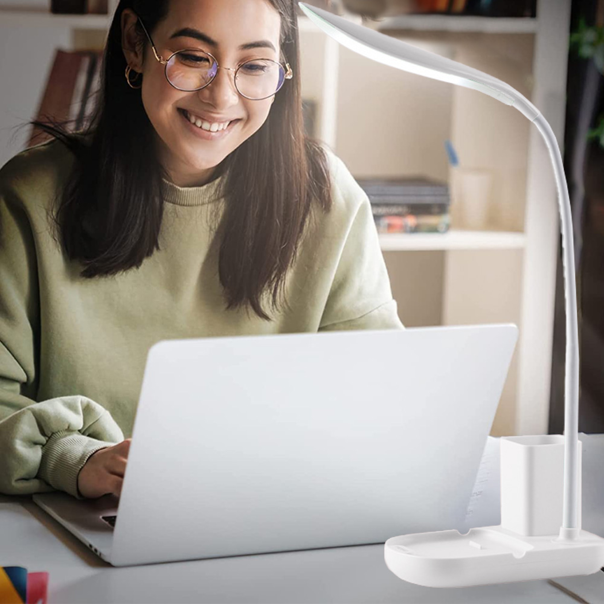 Eine Frau mit Brille lächelt und tippt auf einem Laptop, beleuchtet von einer Schreibtischlampe.