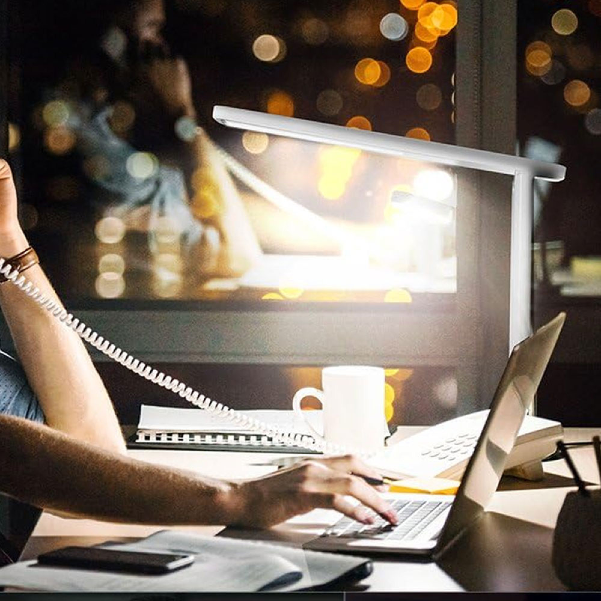 Person tippt auf Laptop, Schreibtisch mit Telefon, Tasse und Lampe. Nachtlichter.
