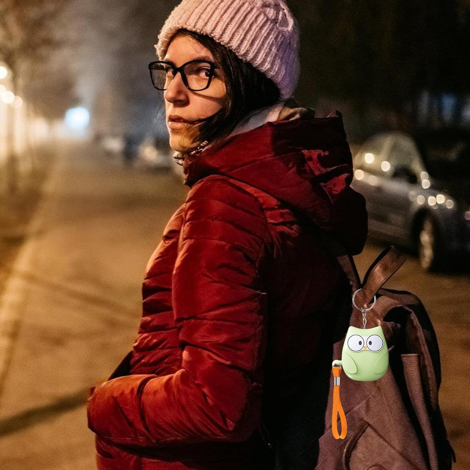 Eine Frau mit Brille und Mütze trägt eine rote Jacke und Rucksack. Ein Eulenanhänger.