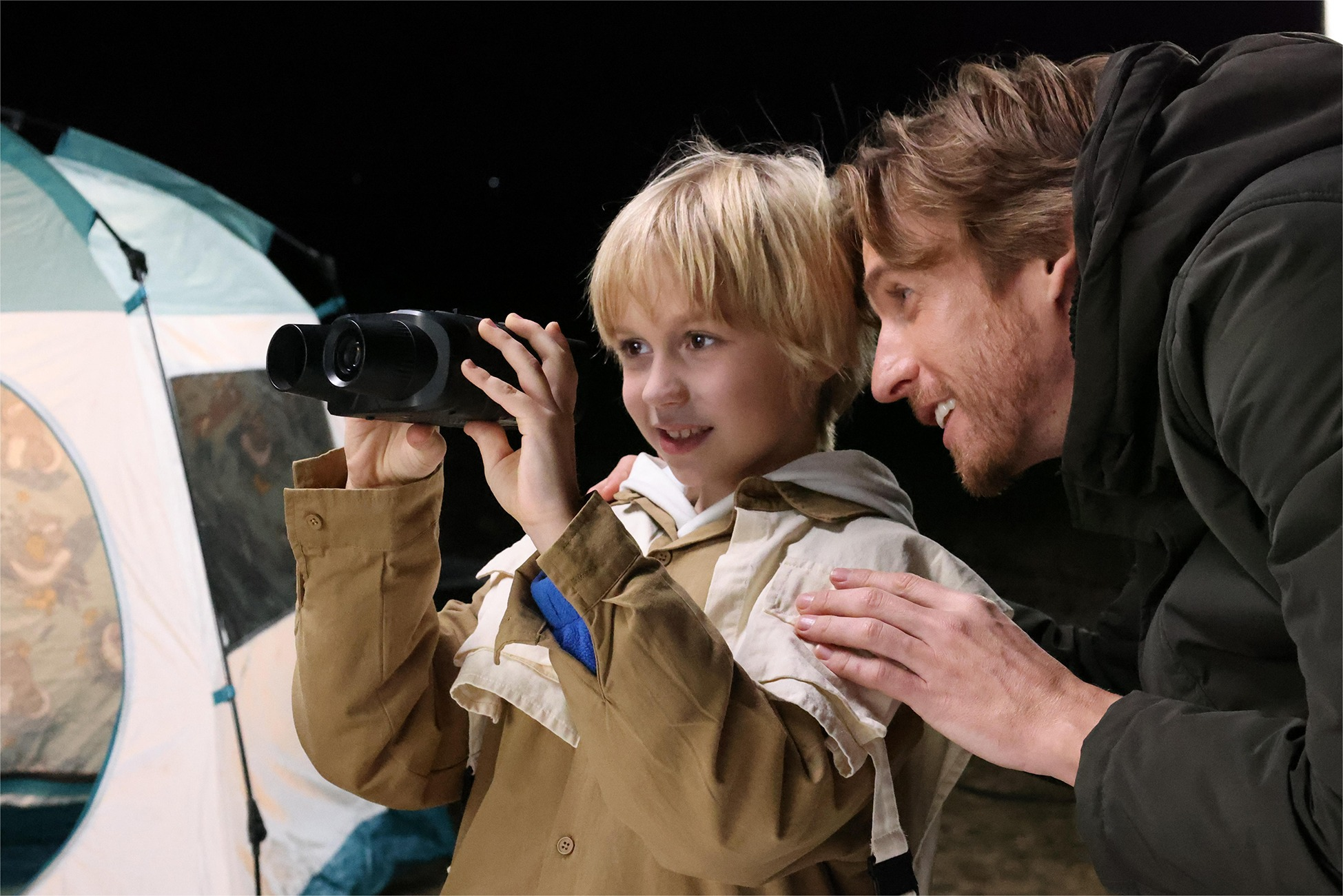 Ein Mann hilft einem Jungen, durch ein Fernglas zu schauen, neben einem Zelt.