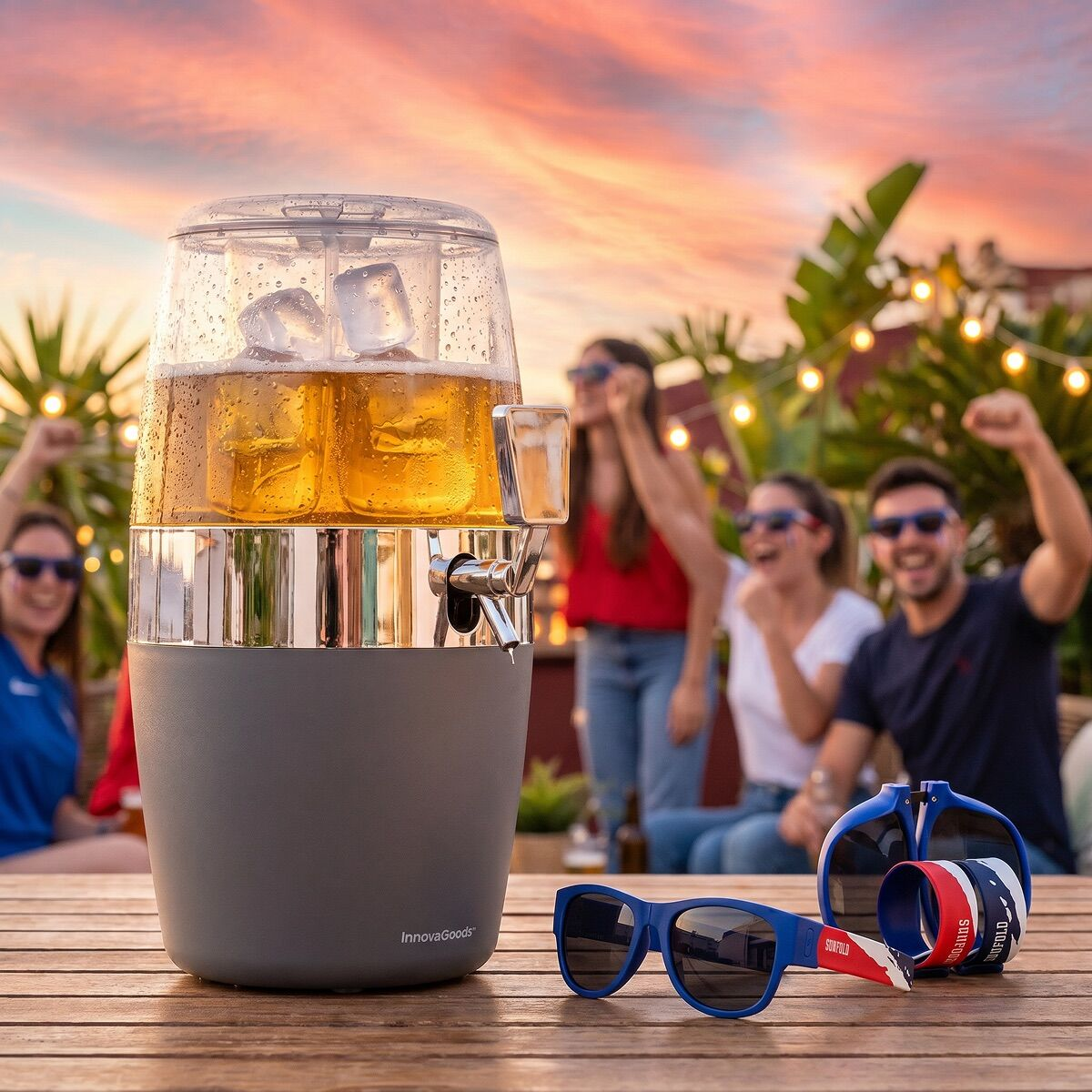 Bierdispenser mit Eis auf einem Tisch, im Hintergrund feiern Menschen bei Sonnenuntergang.